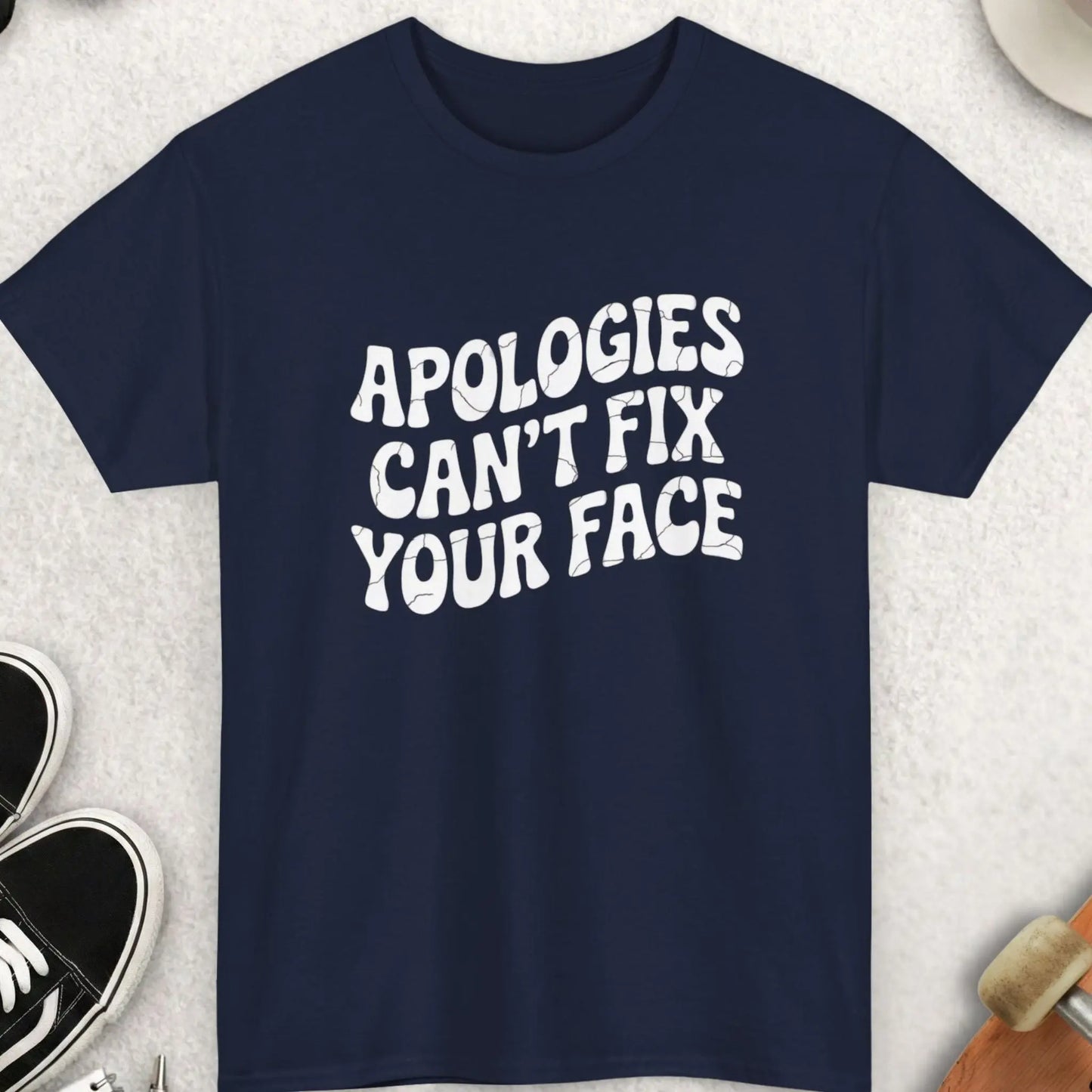 Navy blue t-shirt with 'Apoologies Can't Fix Your Face' text on a light gray surface with accessories.