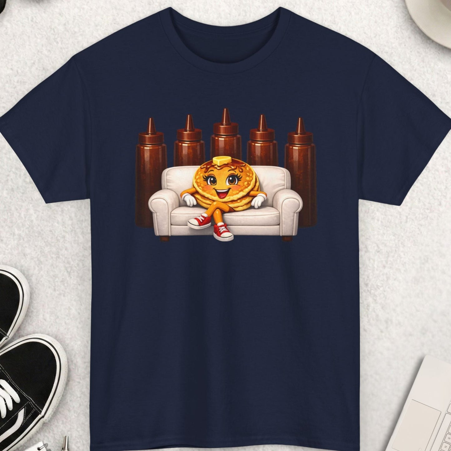 Navy blue t-shirt with a pancake character on a couch graphic, placed on a light gray surface with accessories around.