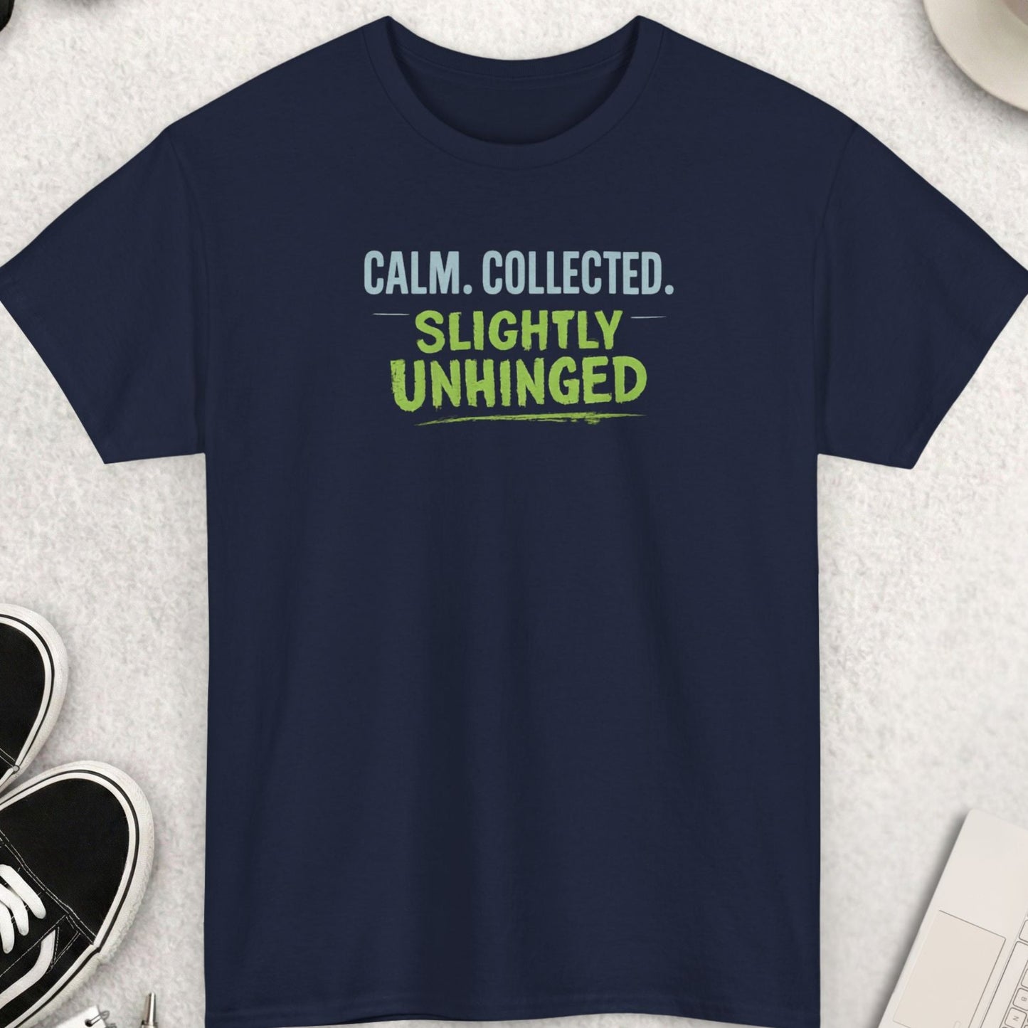 Navy blue t-shirt with text 'CALM. COLLECTED. SLIGHTLY UNHINGED' on a light gray surface with accessories.
