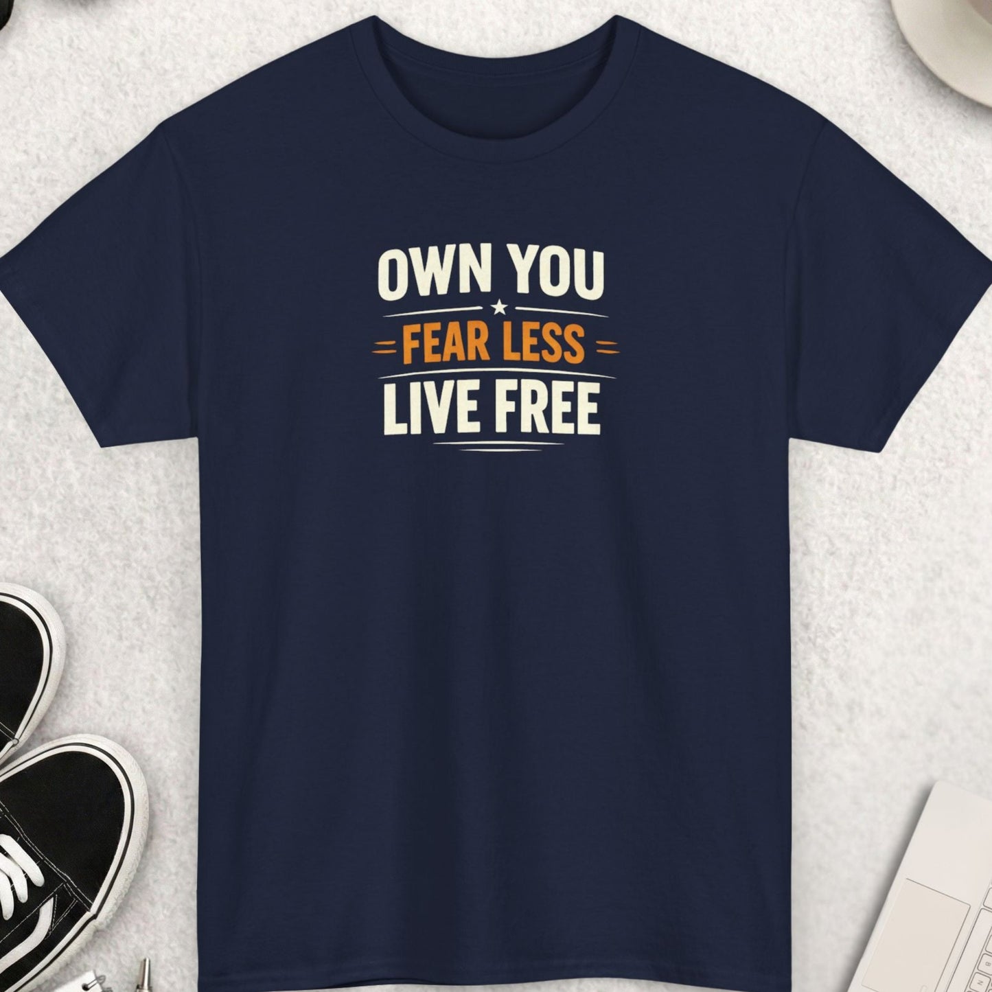 Black t-shirt with OWN YOU. FEAR LESS. LIVE FREE' text on a light gray surface with shoes, sunglasses, and a cup.