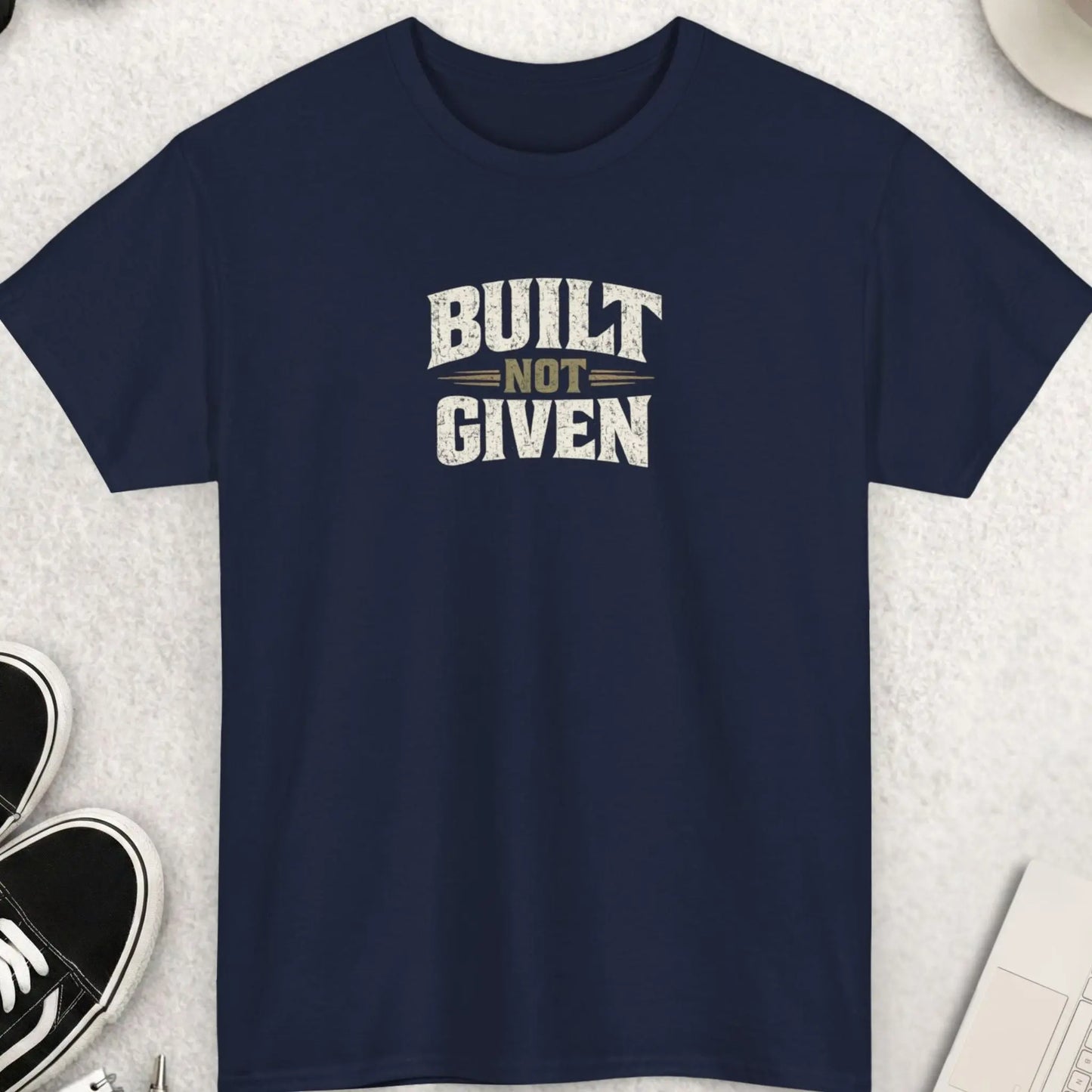Navy blue t-shirt with 'Built Not Given' text on a light gray surface with accessories.