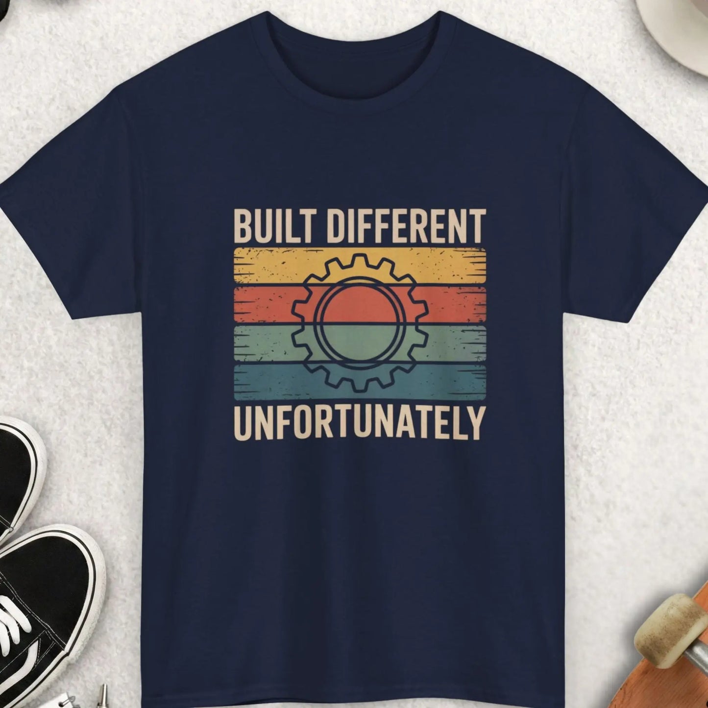 Navy blue t-shirt with 'Built Different, Unfortunately' graphic on a light gray surface with accessories.