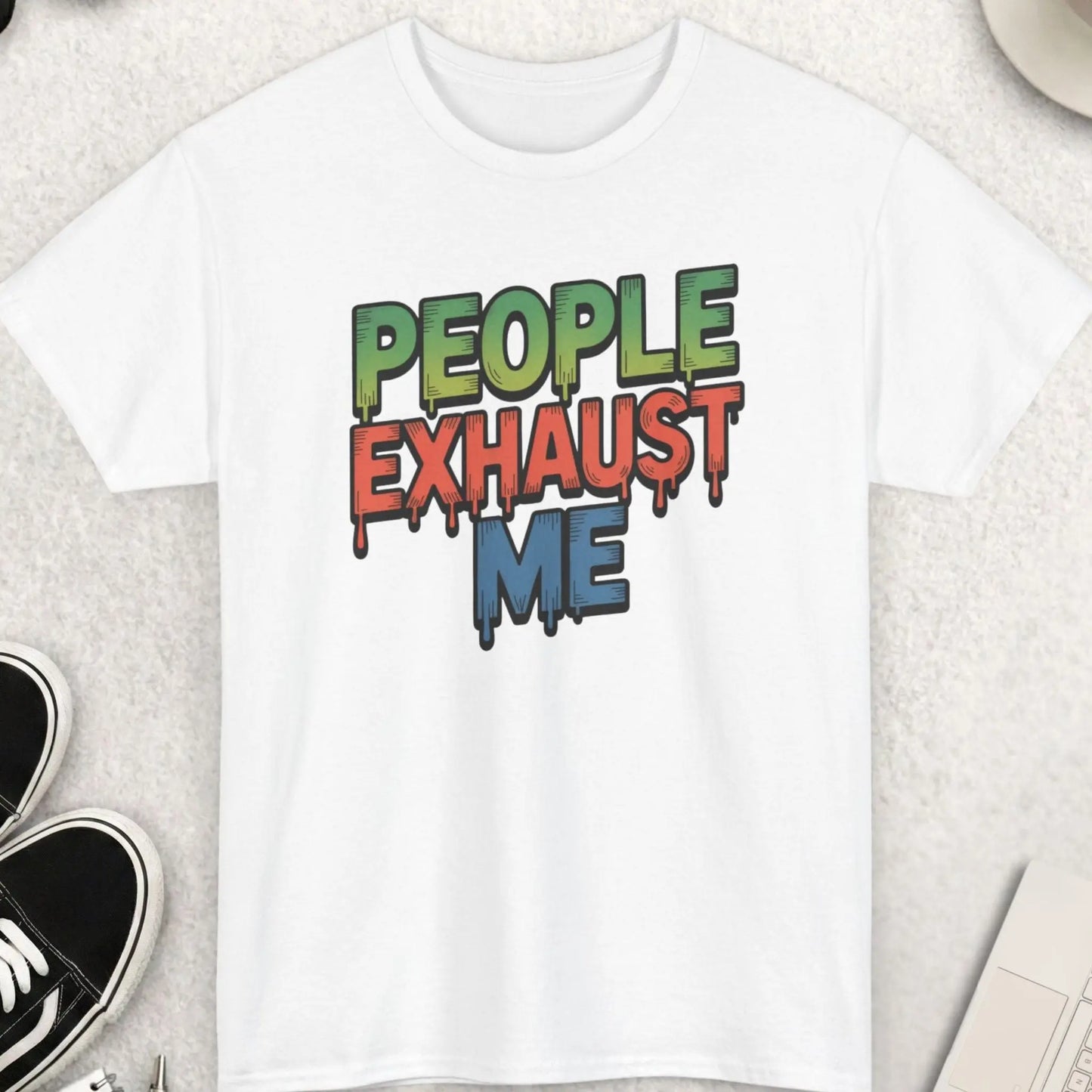 White t-shirt with colorful text 'PEOPLE EXHAUST ME' on a light gray surface with accessories.