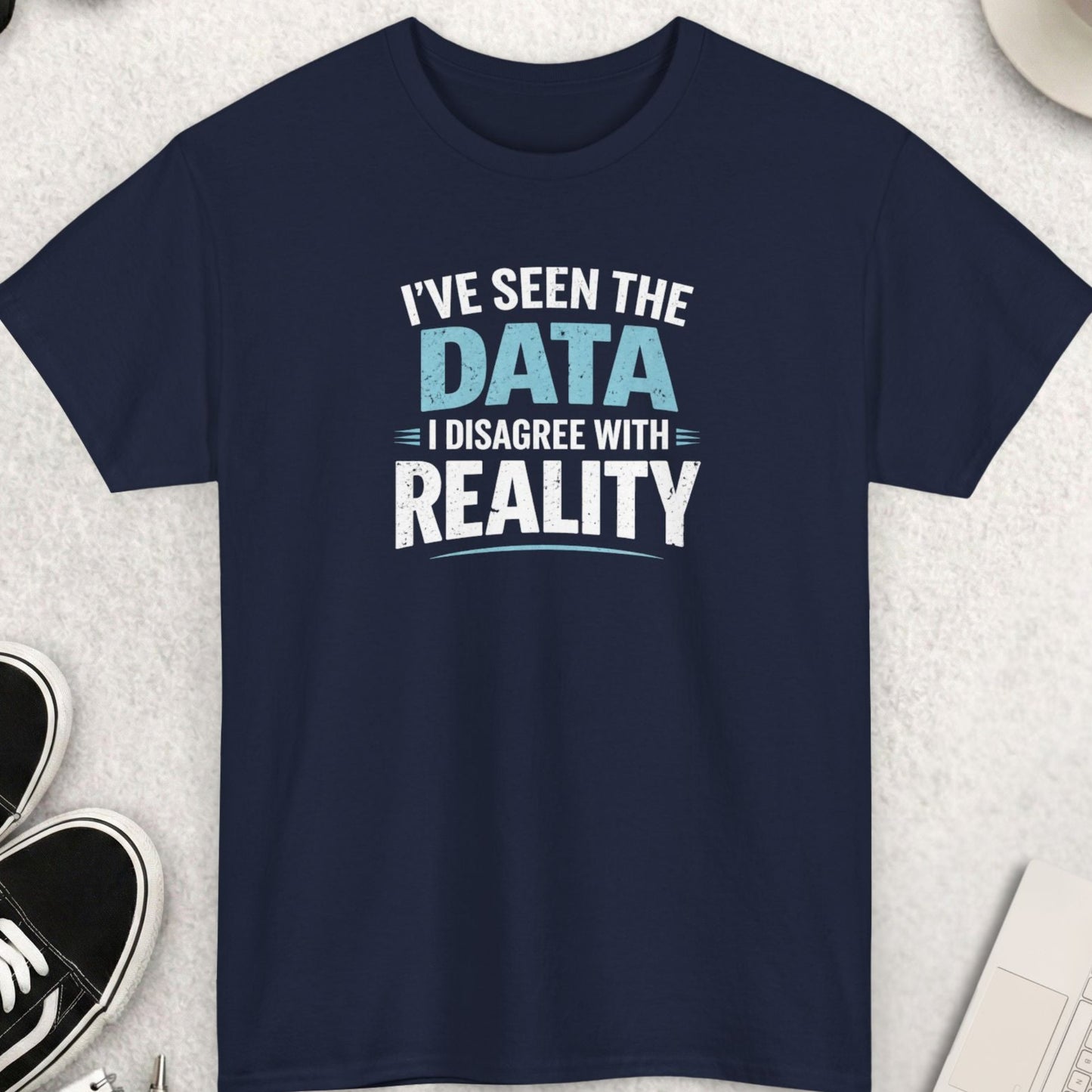 Navy blue t-shirt with text 'I've seen the data I disagree with reality' on a light gray surface with accessories.