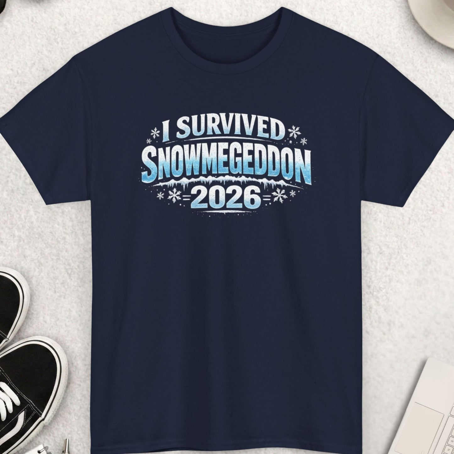 Navy blue t-shirt with 'I Survived Snowmegeddon 2026' text on a light gray surface with shoes, sunglasses, and coffee.