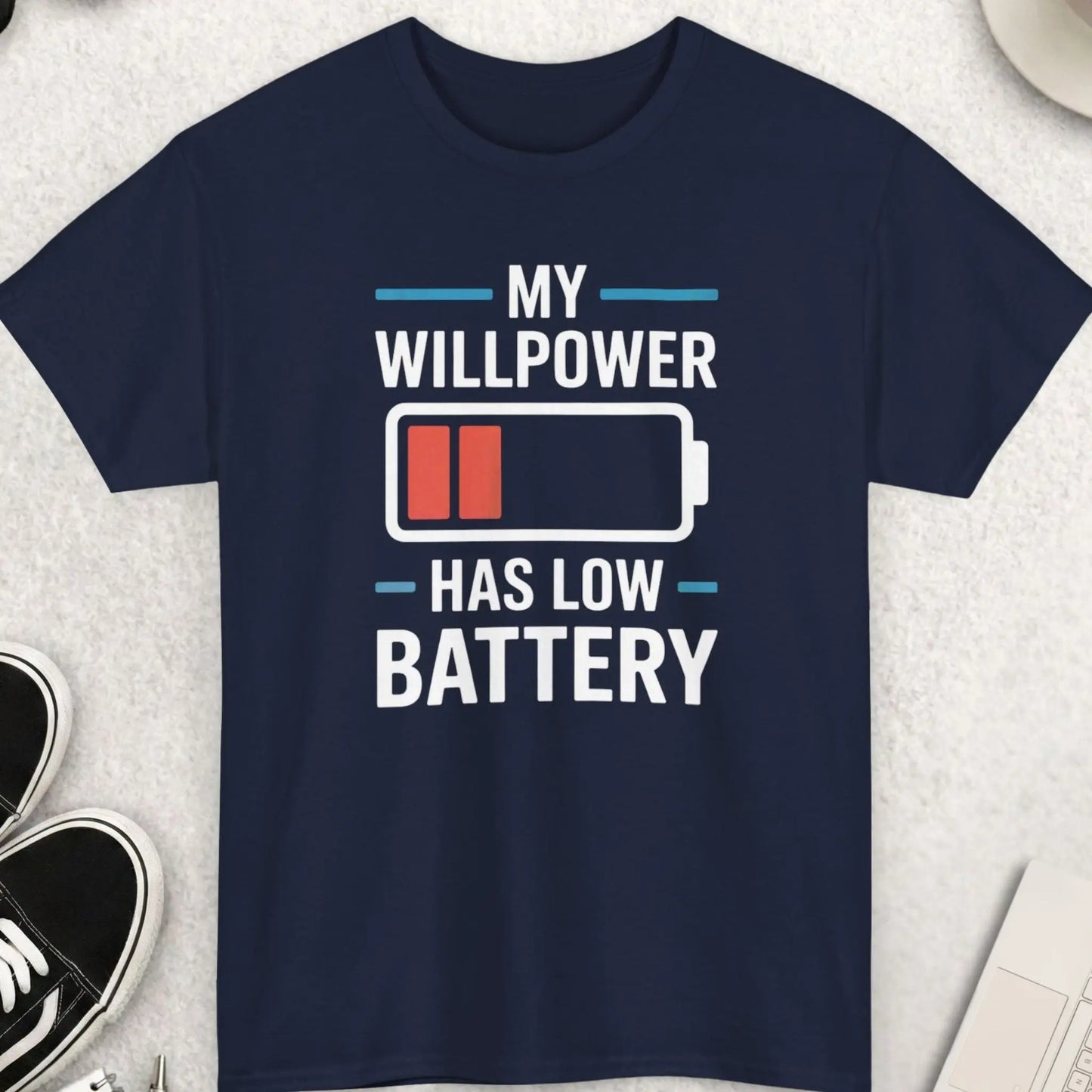 Navy blue t-shirt with 'My willpower has low battery' graphic on a light gray surface with accessories.