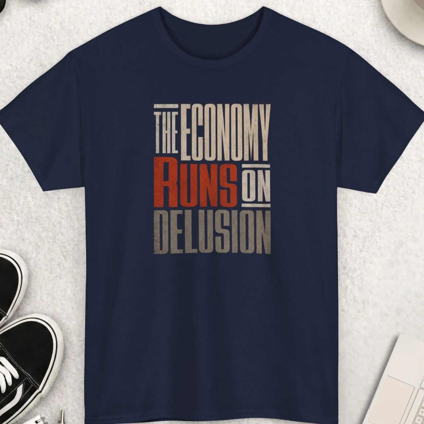 Vintage-style graphic tee featuring the phrase āThe Economy Runs On Delusionā in distressed cream, red, and gray typography on a navy blue shirt with cynical economic commentary vibe.