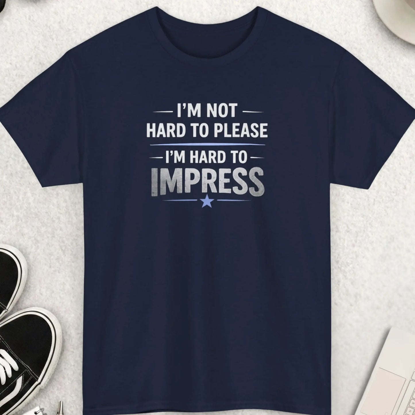 Black t-shirt with 'I’m Not Hard to Please I’m Hard to Impress' text on a light gray surface with shoes, sunglasses, and a cup.