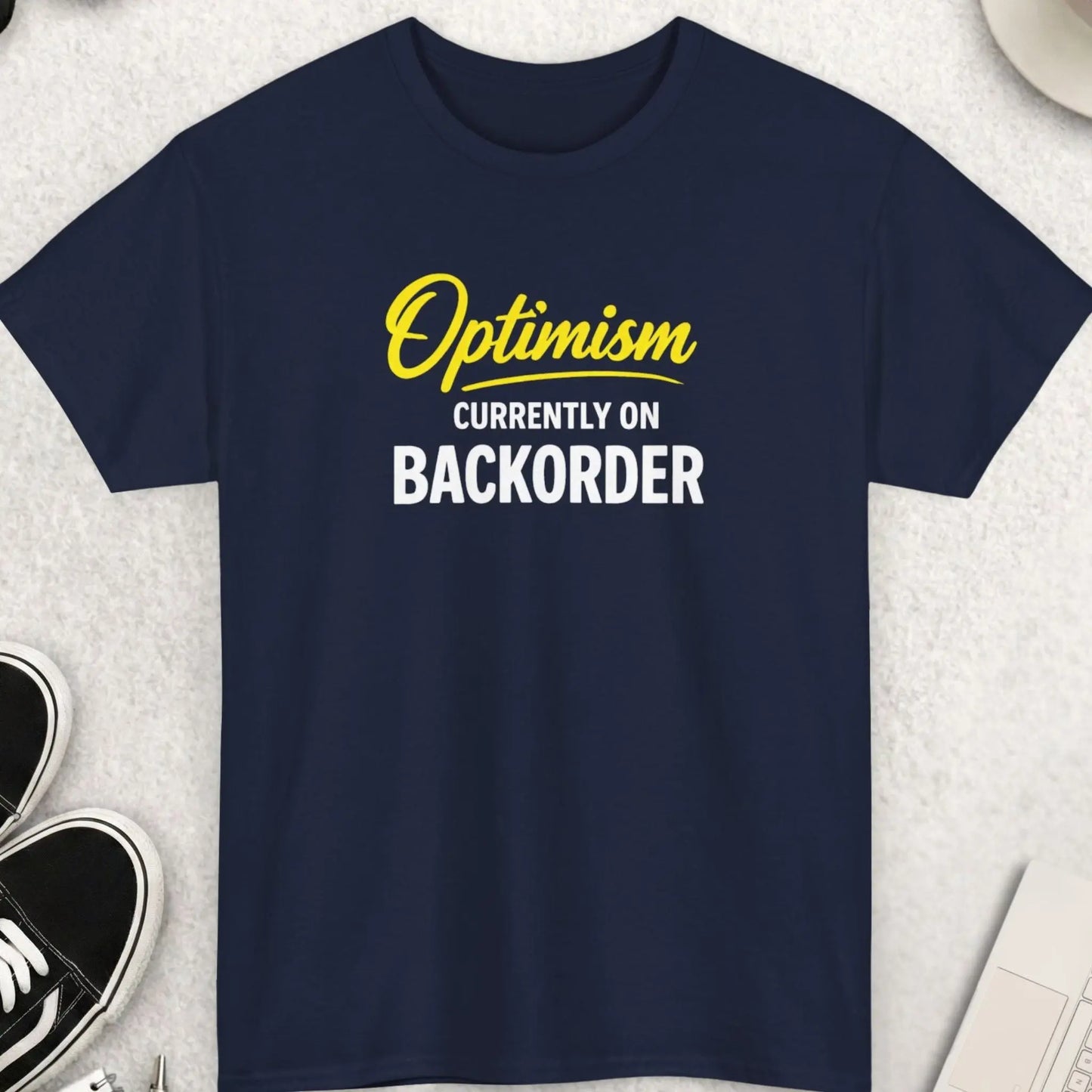 NAVY BLUE t-shirt with 'Black t-shirt with 'Optimism Currently on Back Order'' text on a light gray surface with shoes, sunglasses, and a cup.