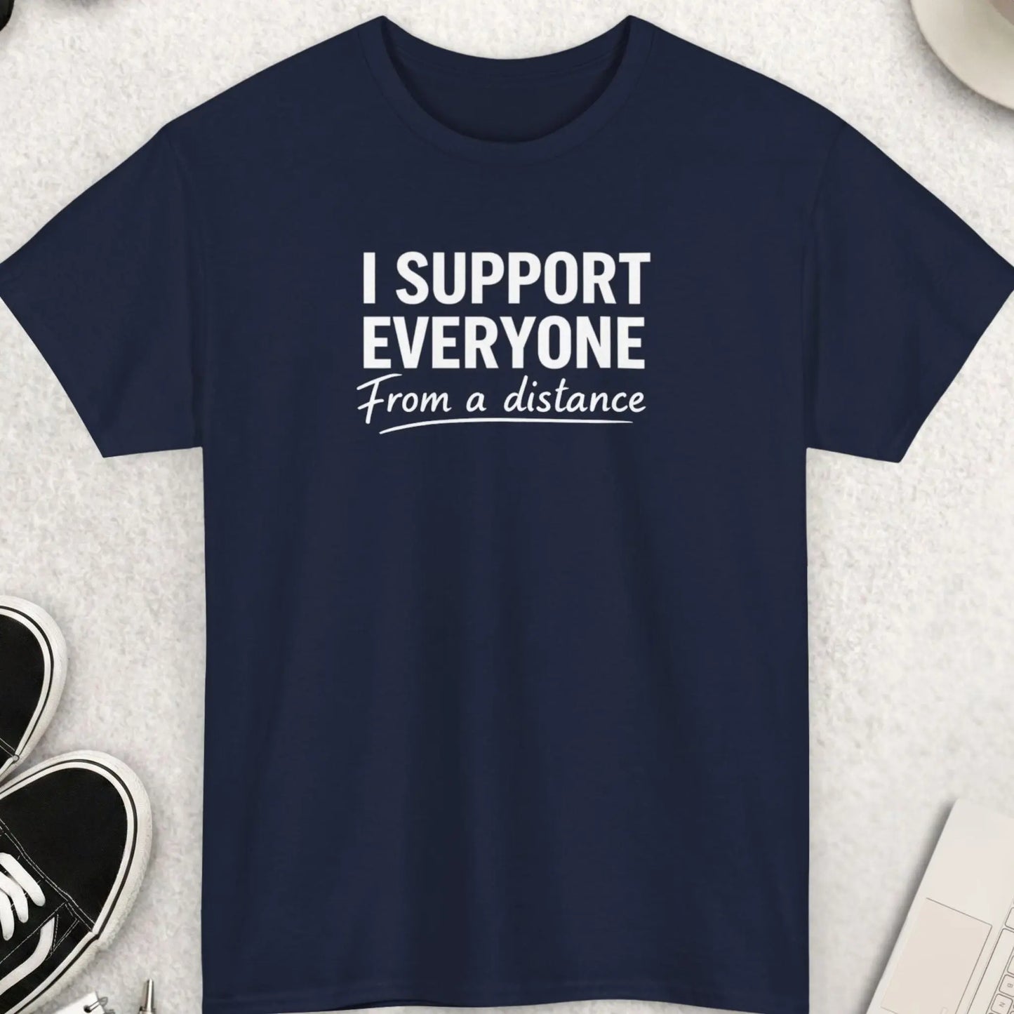 Navy blue t-shirt with 'I Support Everyone From a Distance' text on a light gray surface with shoes, coffee, and accessories.