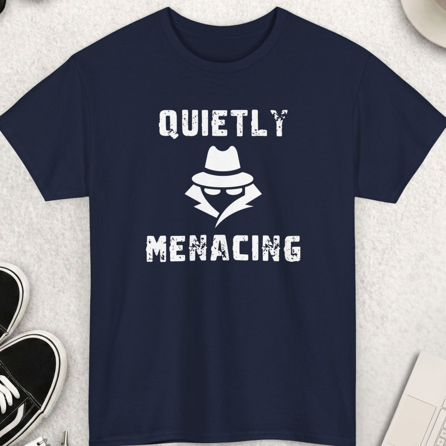 Navy blue t-shirt with 'Quietly Menacing' design on a light gray surface with accessories.