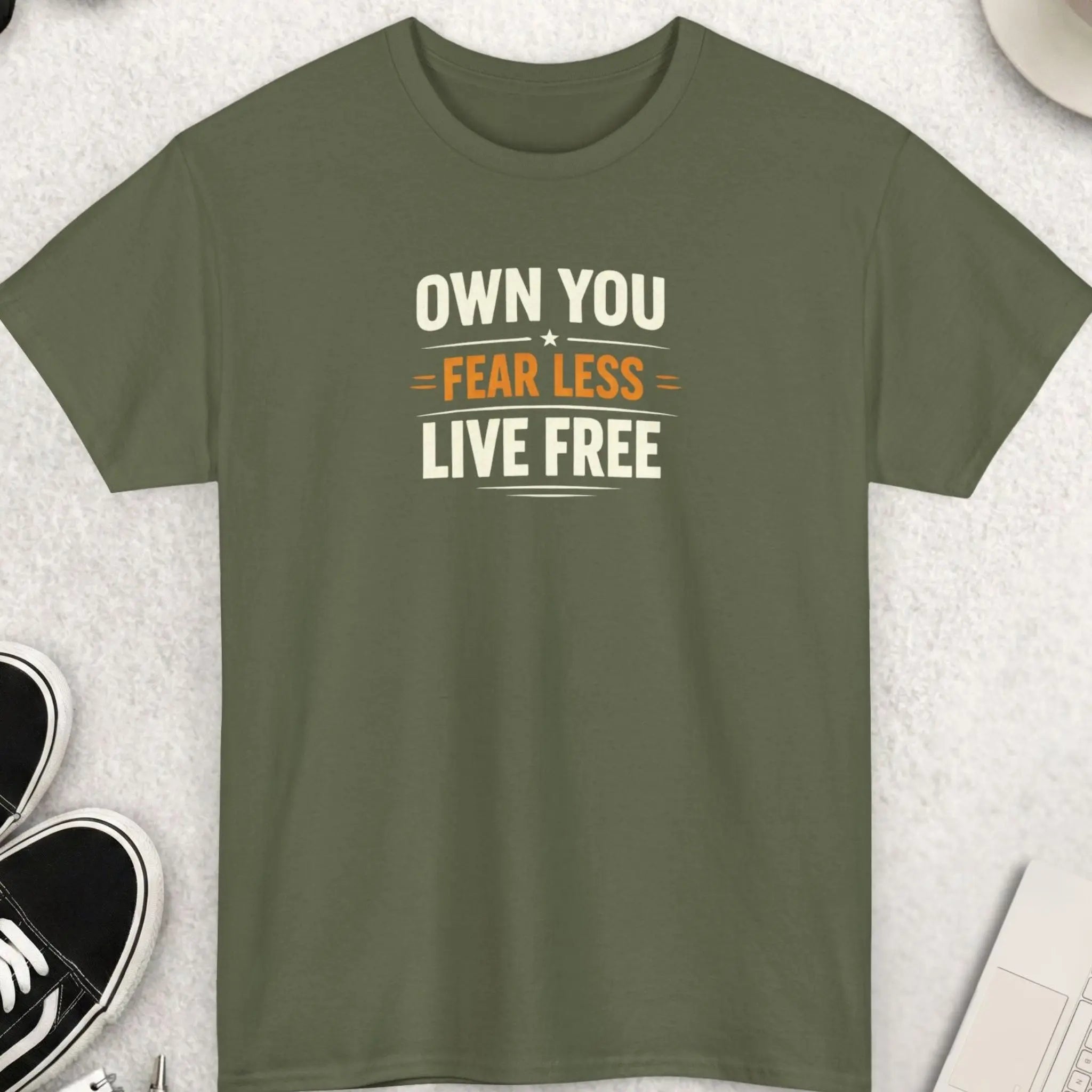 Green t-shirt with 'OWN YOU. FEAR LESS. LIVE FREE' text on a light gray surface with accessories.