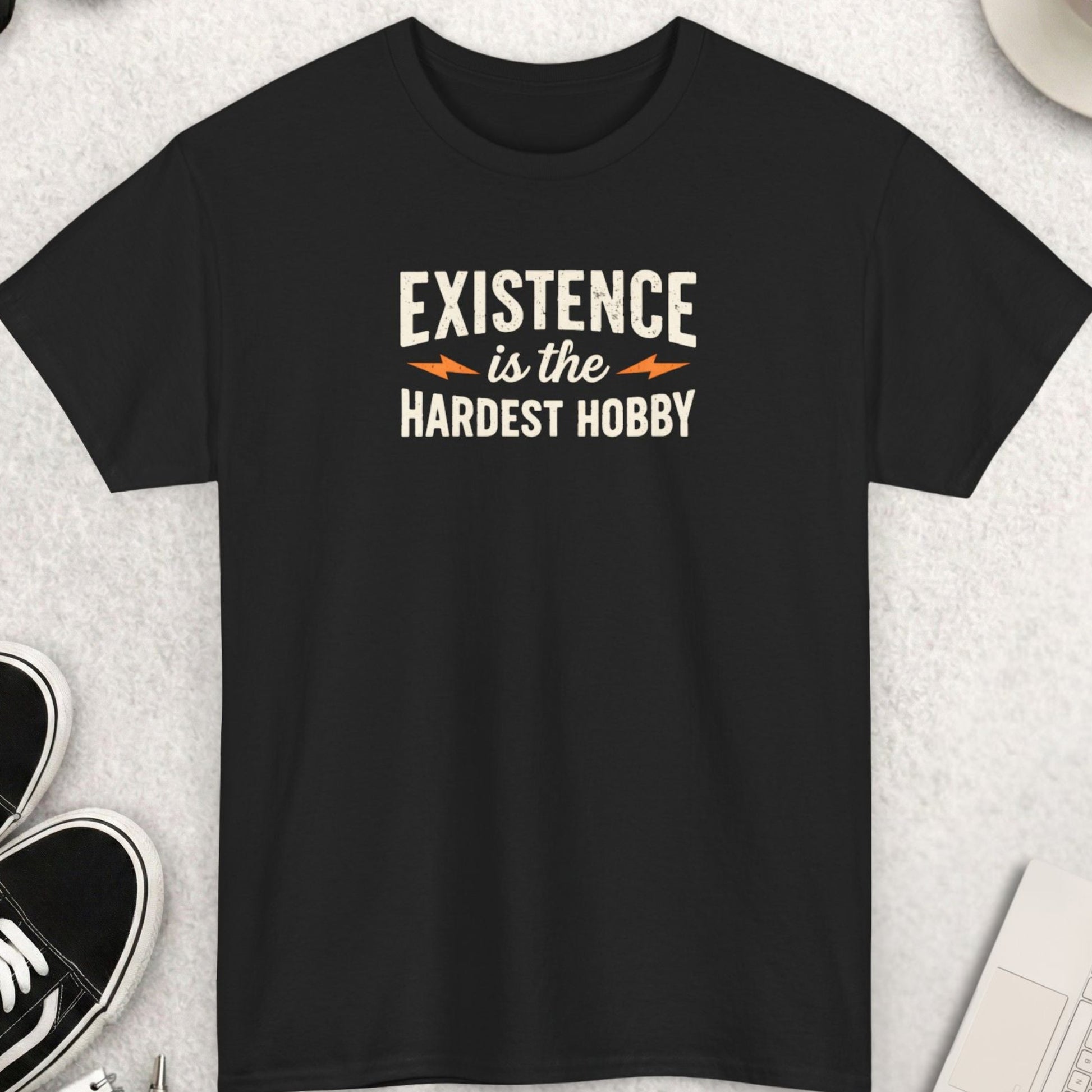 Black t-shirt with 'Existence is the Hardest Hobby' text on a light gray surface with shoes, coffee, and accessories.