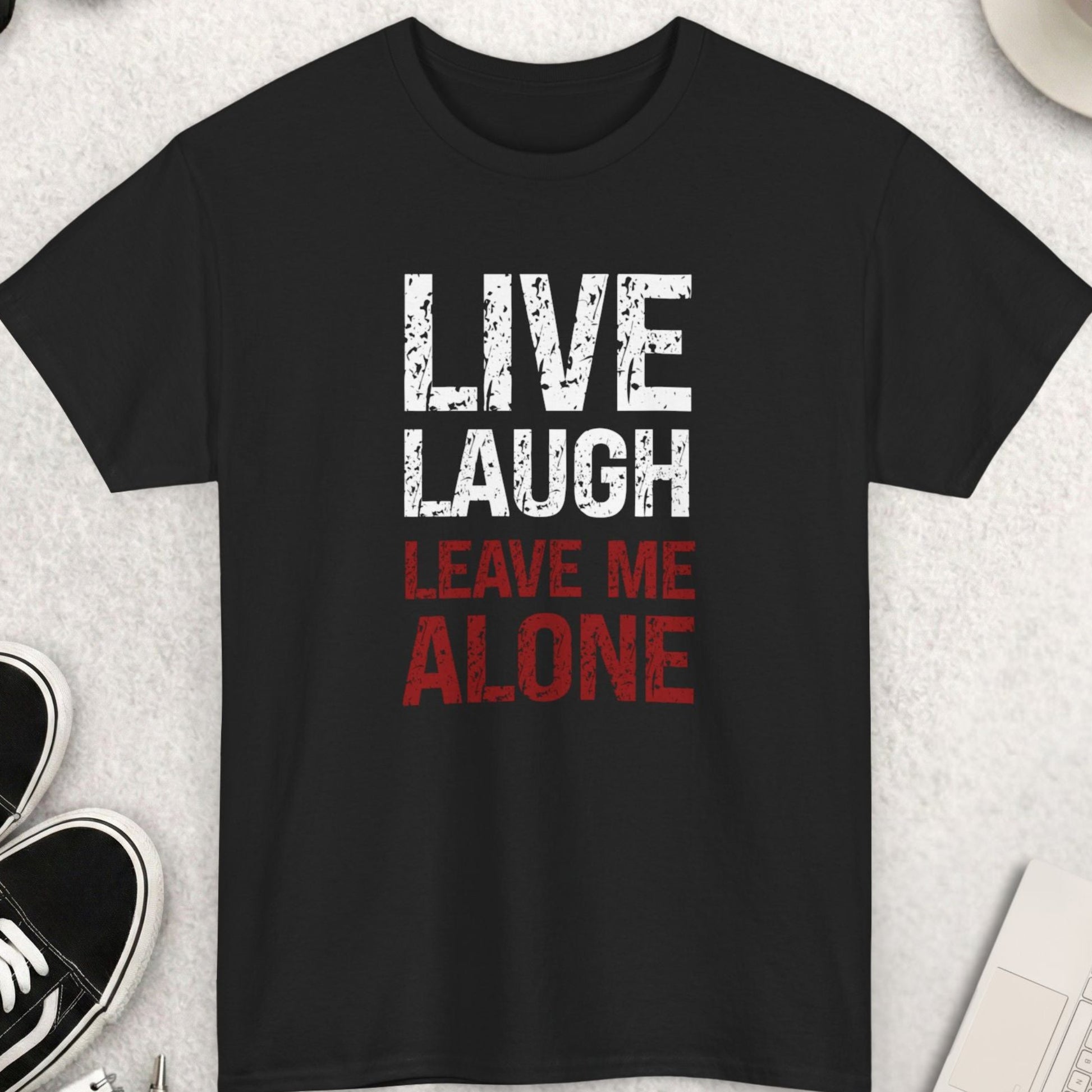 Black t-shirt with 'Live Laugh Leave Me Alone' text on a light gray surface with shoes, coffee, and sunglasses.