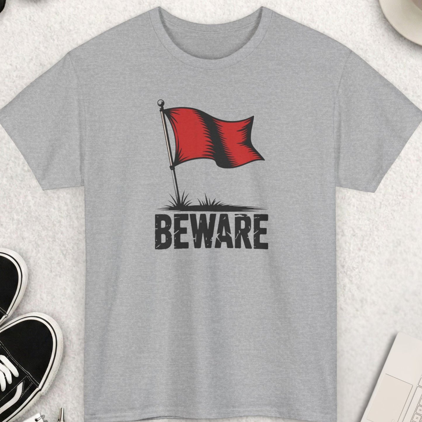 Gray t-shirt with a red flag and 'Beware' text on a light gray surface with shoes, sunglasses, and a cup of coffee.