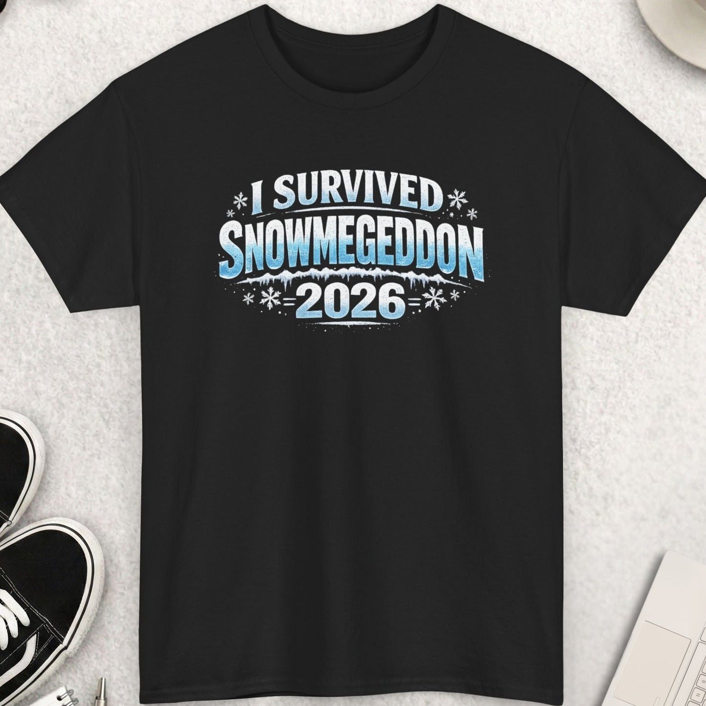 Black t-shirt with 'I Survived Snowmegeddon 2026' text on a light gray surface with shoes, coffee, and sunglasses.