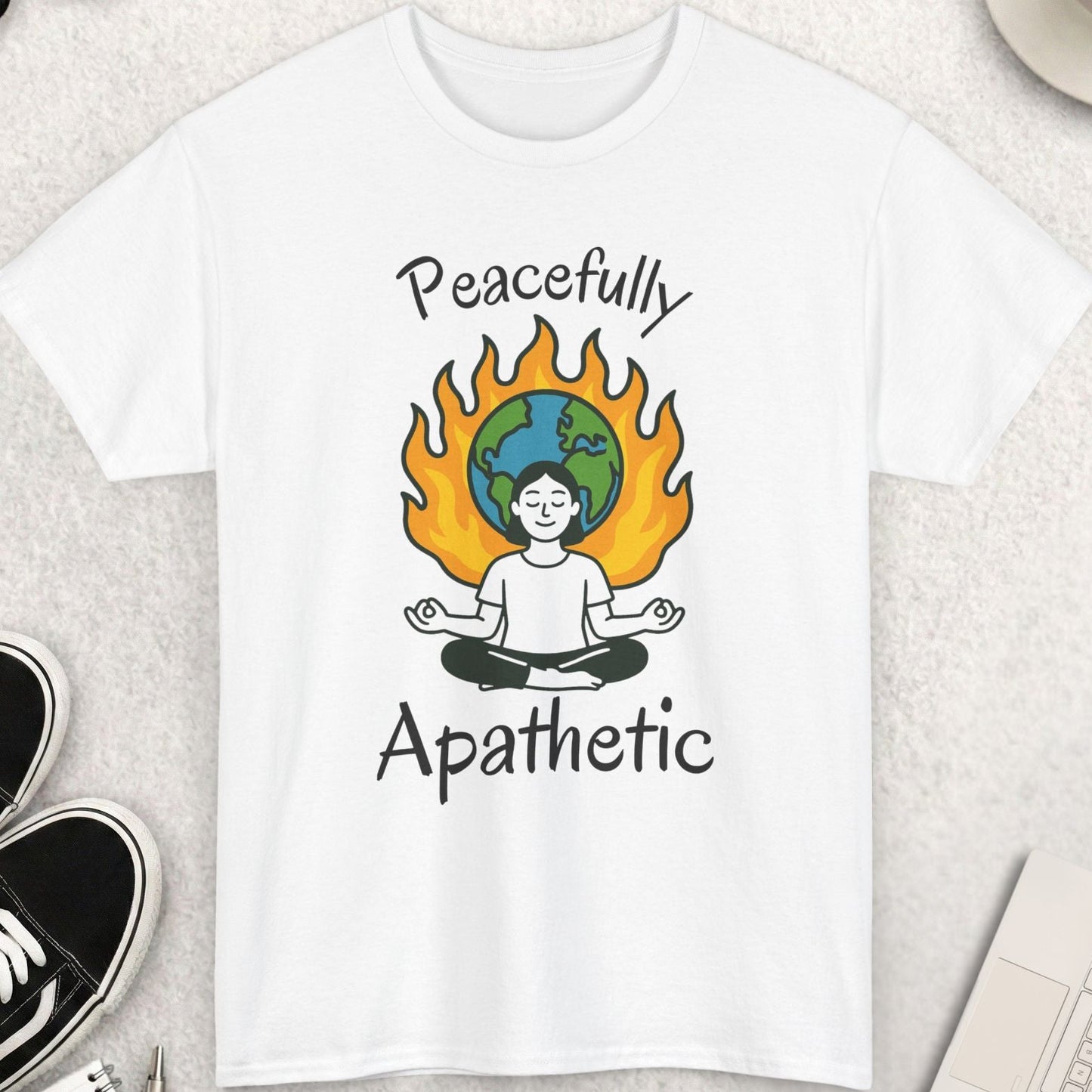White t-shirt with 'Peacefully Apathetic' design featuring a person meditating with a flame and world map, placed on a light surface with accessories around.