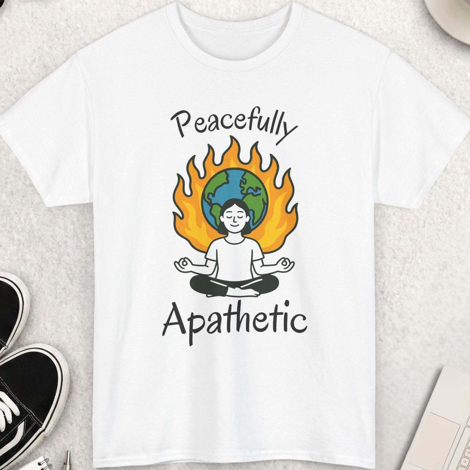 White t-shirt with 'Peacefully Apathetic' design featuring a person meditating with a flame and world map, placed on a light surface with accessories around.