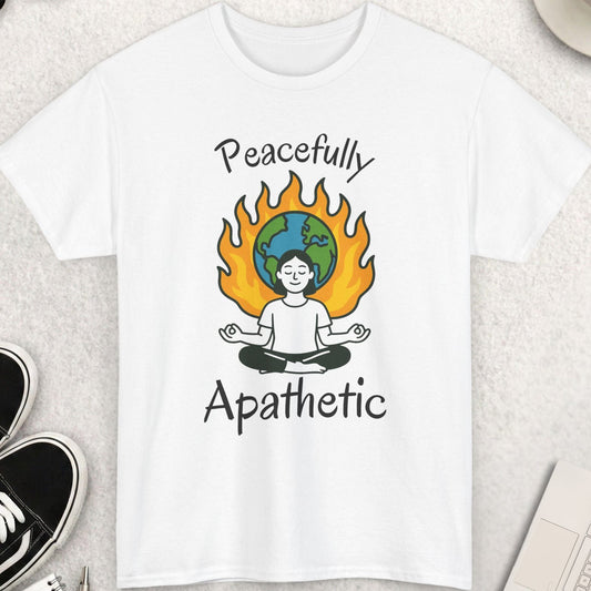 White t-shirt with 'Peacefully Apathetic' design featuring a person meditating with a flame and world map, placed on a light surface with accessories around.