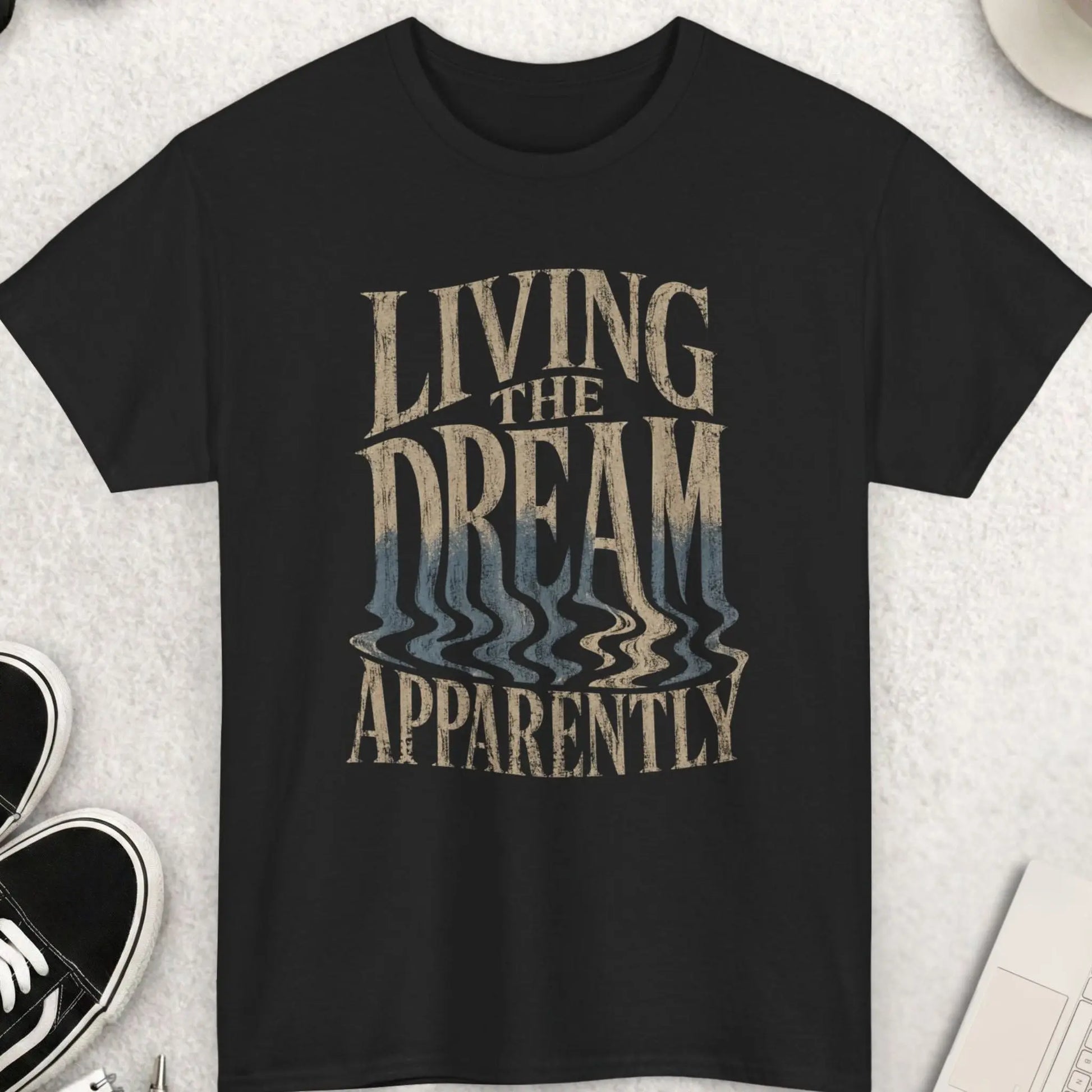 Black distressed typography graphic t-shirt featuring the phrase “Living The Dream Apparently” with warped melting cream and faded blue lettering in a dark humor grunge streetwear design by Mayhem Haus.