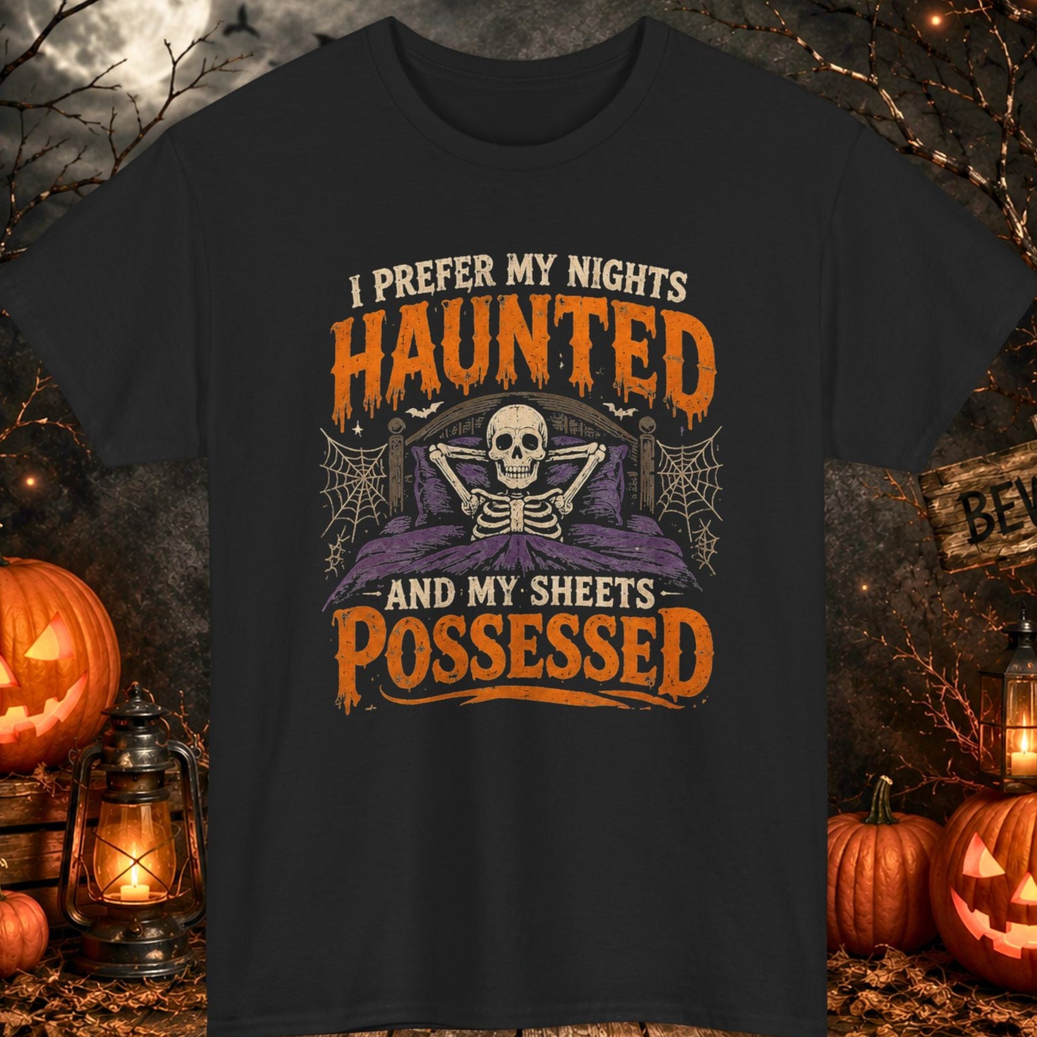 Mayhem Haus A black graphic t-shirt featuring a Halloween design with a skull, spiderwebs, and orange text reading 'I PREFER MY NIGHTS HAUNTED AND MY SHEETS POSSESSED'. The shirt is displayed in a spooky outdoor scene with pumpkins, lanterns, bare trees, and a full moon.