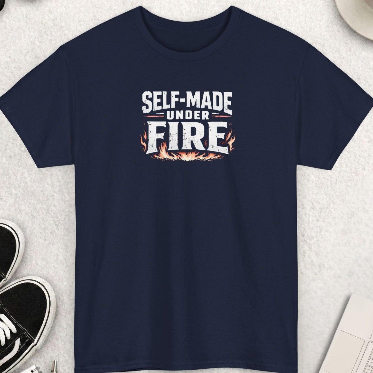 Navy blue t-shirt with 'Self-Made Under Fire' text on a light gray surface with accessories.