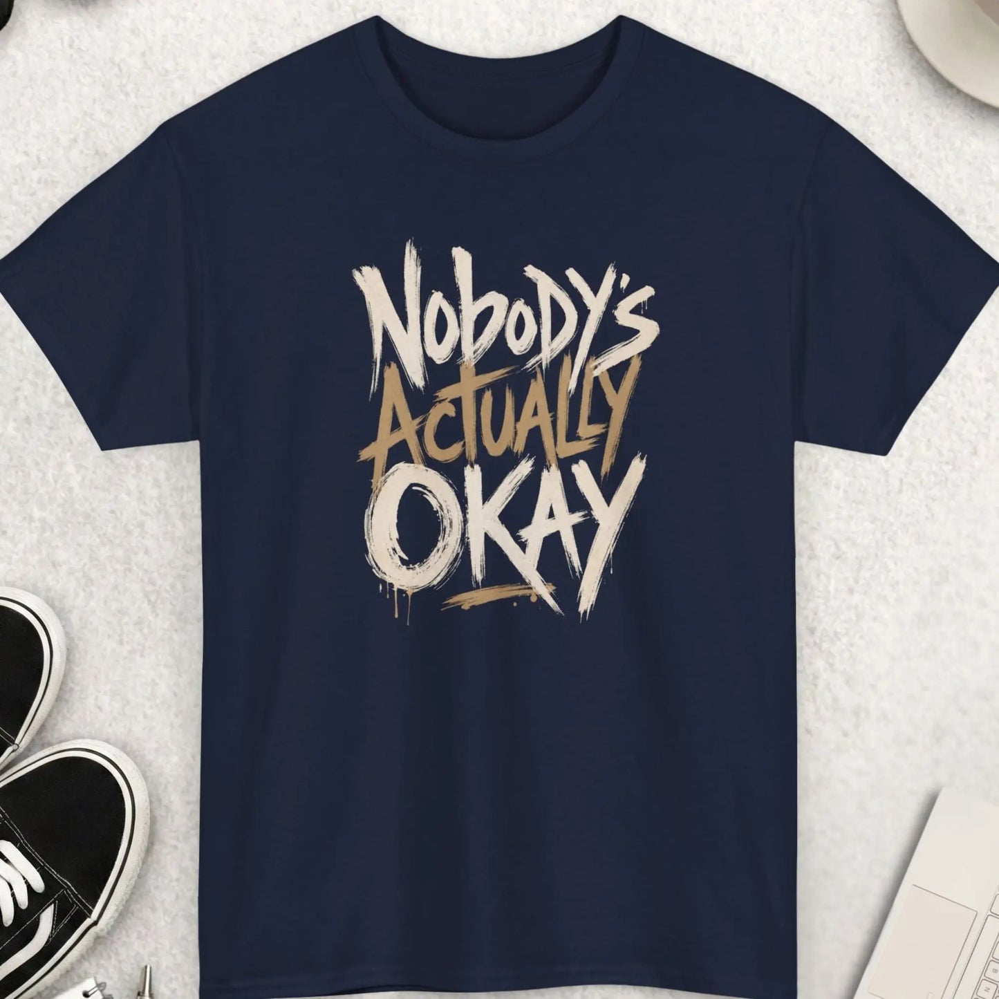 Navy blue t-shirt with aggressive brush-style typography displaying the phrase “Nobody’s Actually Okay” in a distressed modern layout.