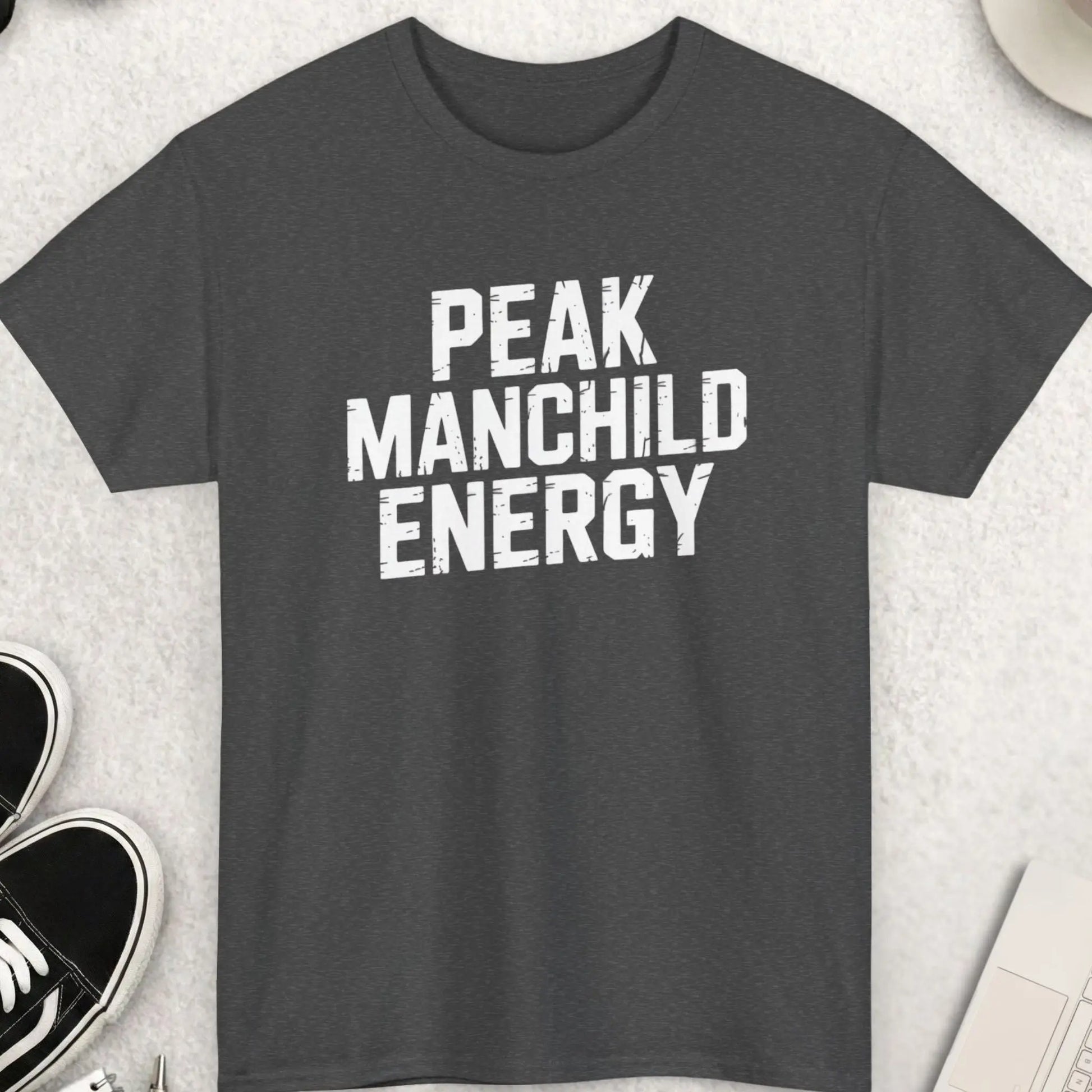 Dark gray t-shirt with 'Peak Manchild Energy' text on a light gray surface with shoes, coffee, and sunglasses.