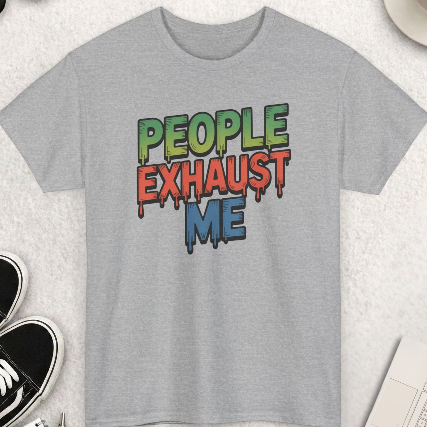 Gray t-shirt with colorful text 'PEOPLE EXHAUST ME' on a light surface with shoes, coffee, and a keyboard.