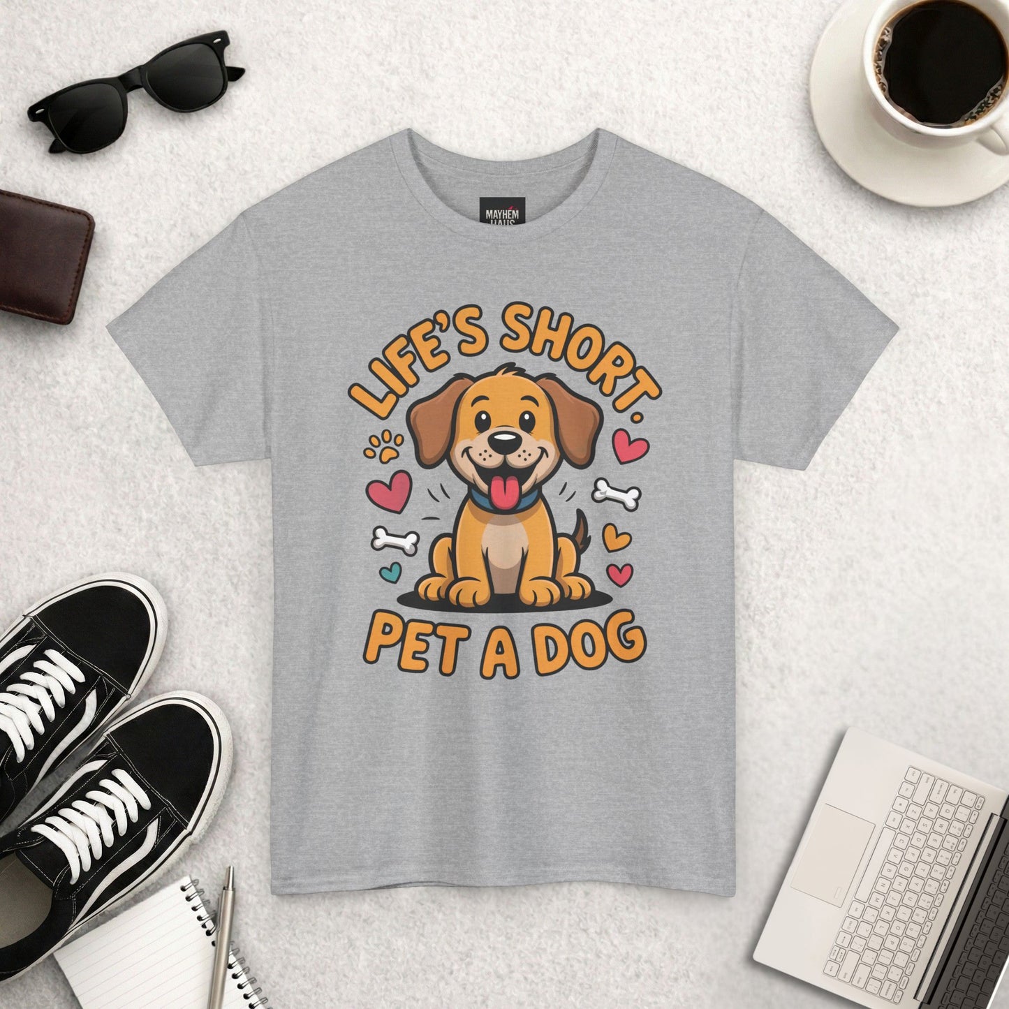 Gray t-shirt with a dog graphic and text 'Life's Short, Pet a Dog' on a textured surface with shoes, coffee, and accessories.