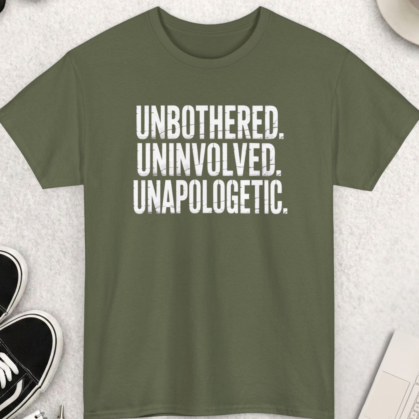 Green t-shirt with 'UNBOtherED. UNINVOLVED. UNAPOLOgetic.' text on a light gray surface with shoes, sunglasses, and coffee.
