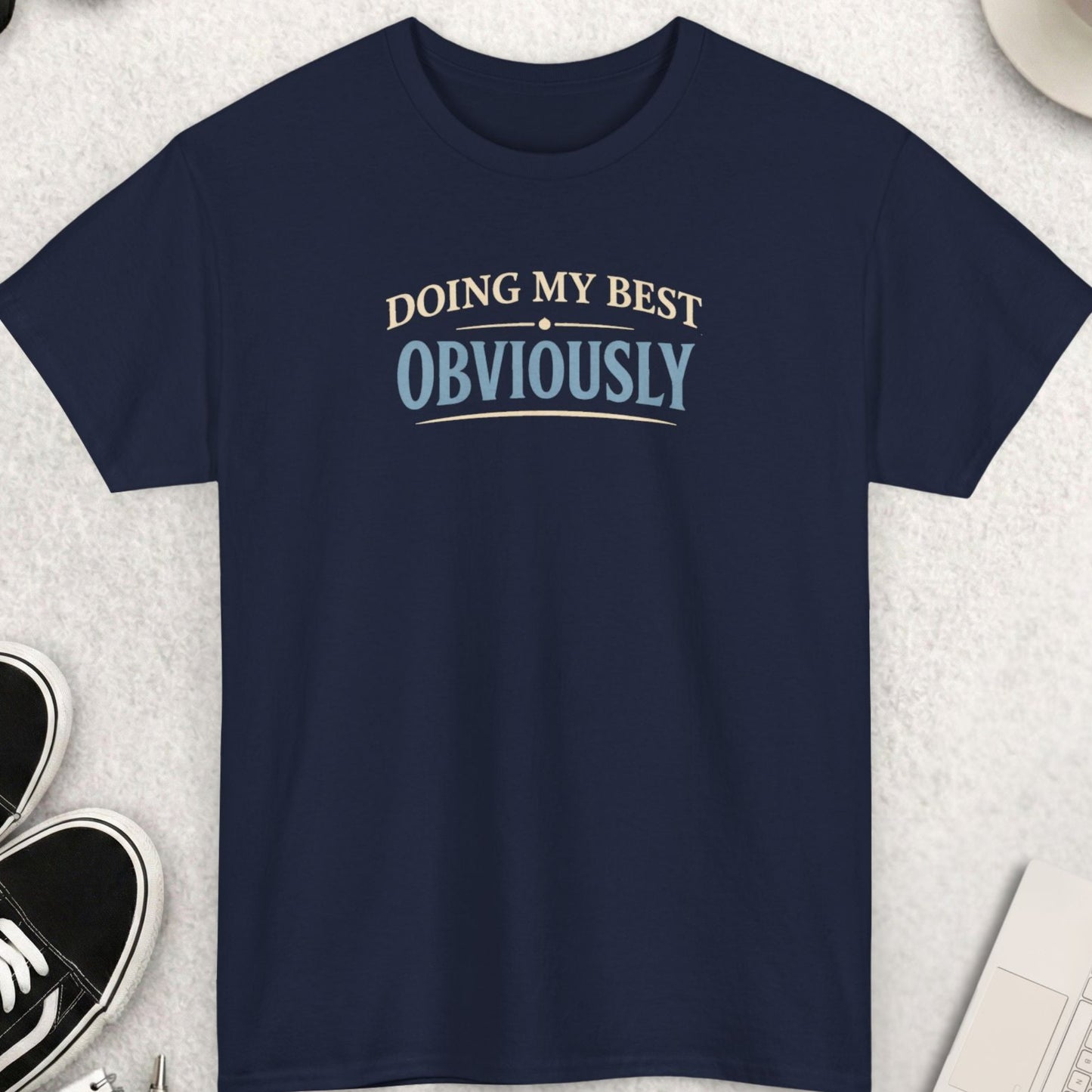 Navy blue t-shirt with 'Doing My Best Obviously' text on a light gray surface with accessories.