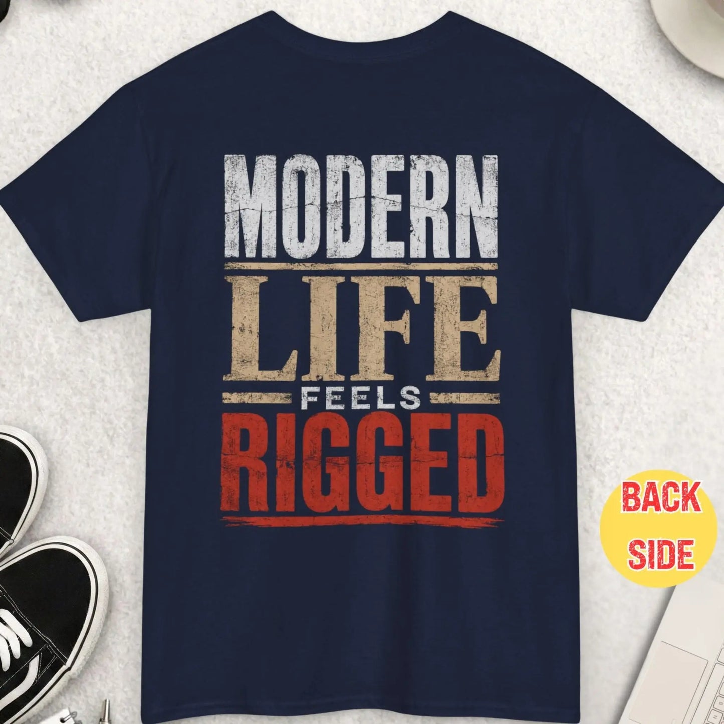Navy blue t-shirt with distressed typography displaying the phrase “Modern Life Feels Rigged” in a worn vintage-style layout with red accents.