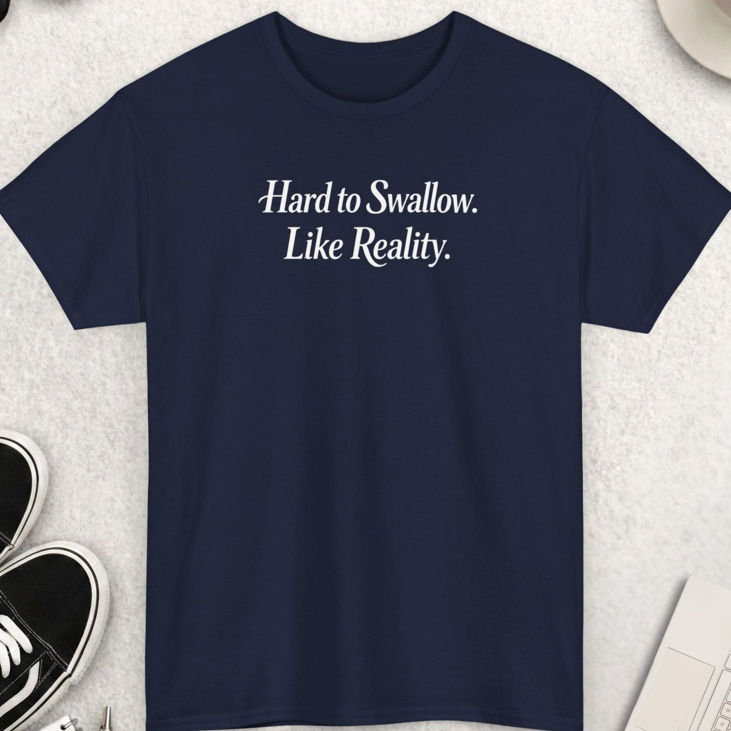Navy blue t-shirt with 'Hard to Swallow. Like Reality.' text on a light gray surface with shoes, coffee, and accessories.