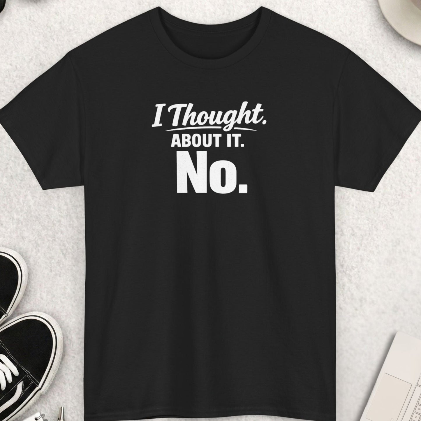 Black t-shirt with 'I Thought About It. No.' text on a light gray surface with shoes, coffee, and accessories.