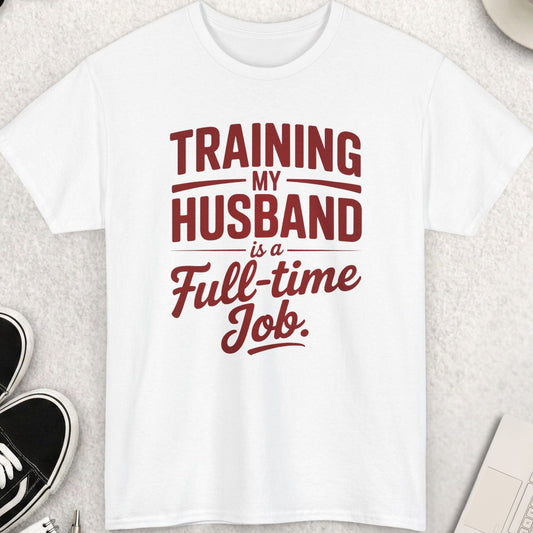 White t-shirt with red text 'TRAINING MY HUSBAND IS A FULL-TIME JOB' on a light gray surface with accessories.