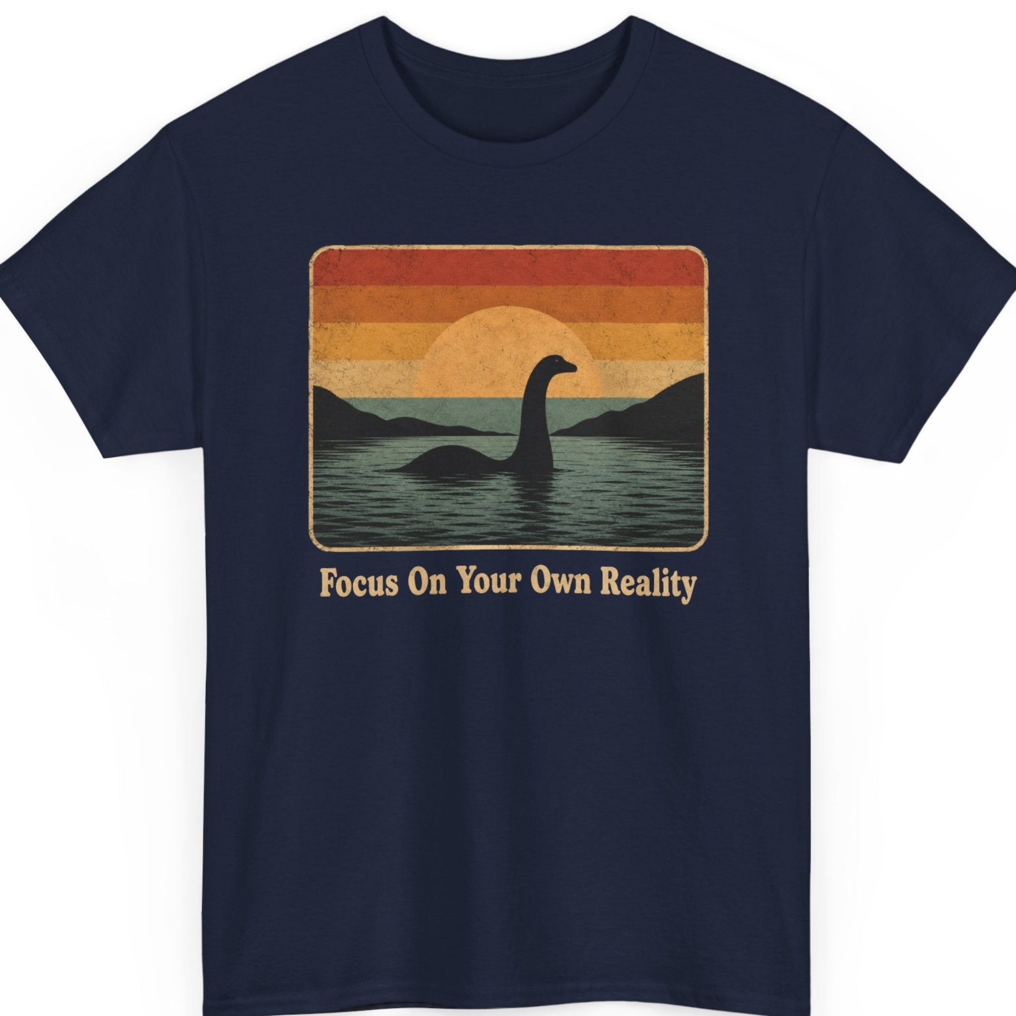 Mayhem Haus cryptid t-shirt on a navy blue shirt featuring a retro sunset landscape, calm lake, mountain silhouettes, and the iconic Loch Ness Monster with the quote "Focus On Your Own Reality."