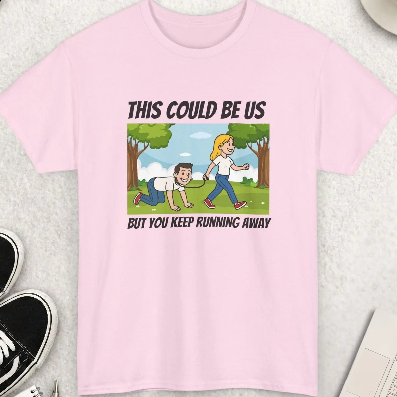 Pink t-shirt with a graphic and text on a light gray surface with accessories around.