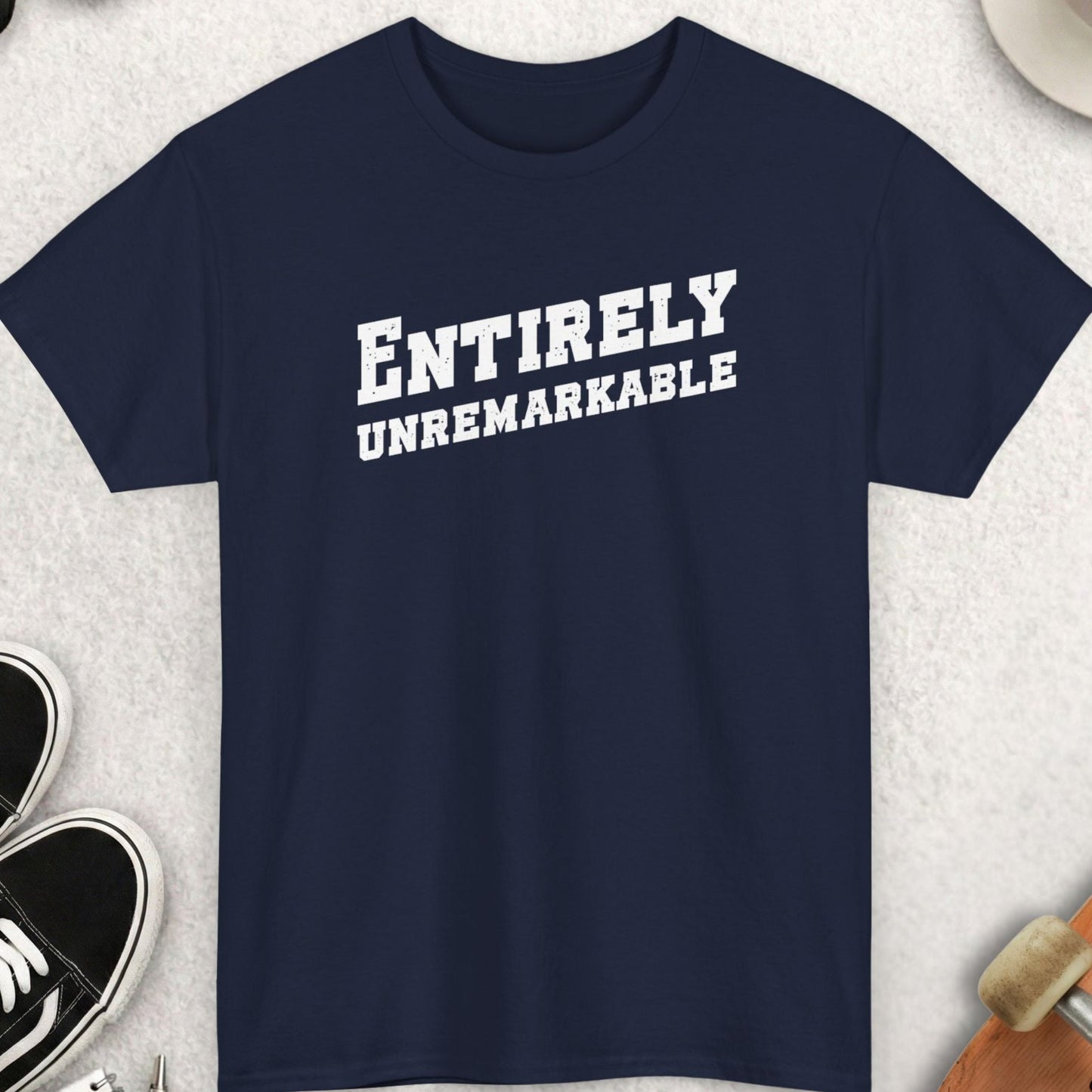 Navy blue t-shirt with 'ENTIRELY UNREMARKABLE' text, surrounded by casual items on a light surface.