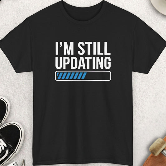 Black t-shirt with 'I'm Still Updating' text on a light gray surface with accessories.