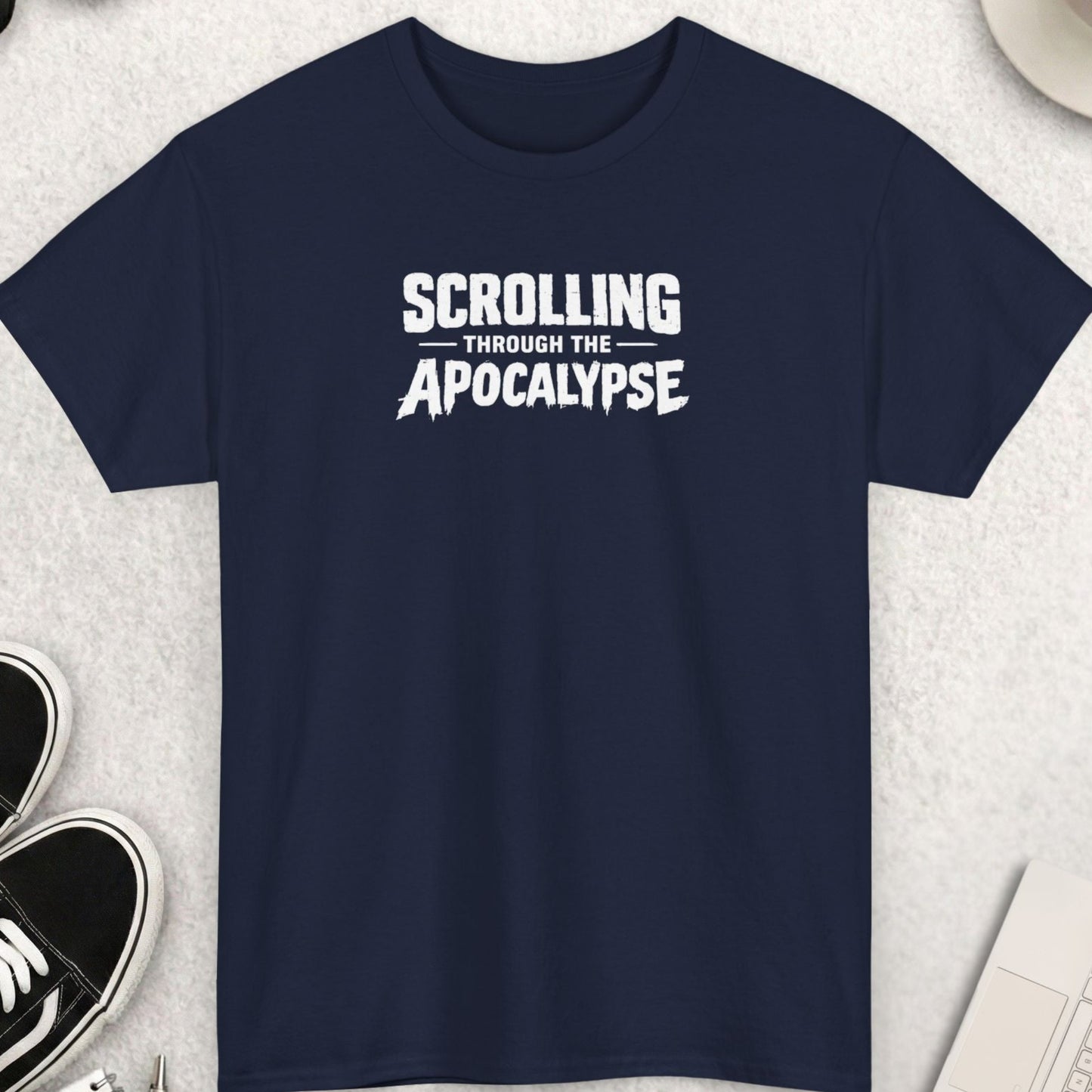 Black t-shirt with 'Scrolling Through the Apocalypse' text on a light gray surface with shoes, sunglasses, and a cup of coffee.