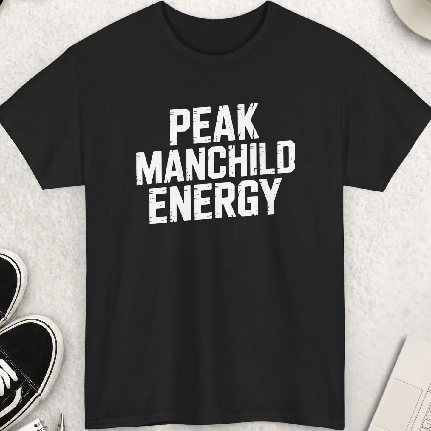 Black t-shirt with 'PEAK MANCHILD ENERGY' text on a light gray surface with accessories.