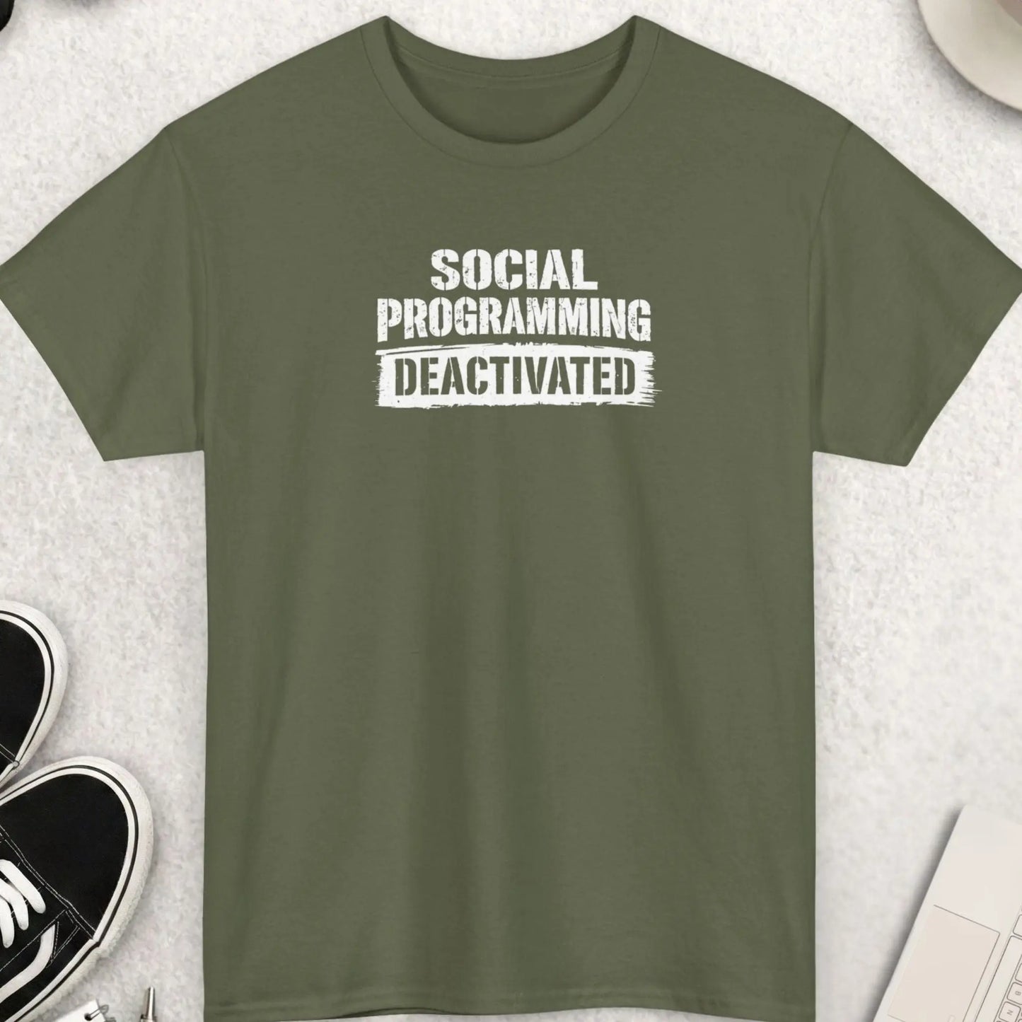 Green t-shirt with 'SOCIAL PROGRAMMING DEACTIVATED' text on a light gray surface with shoes, coffee, and accessories.