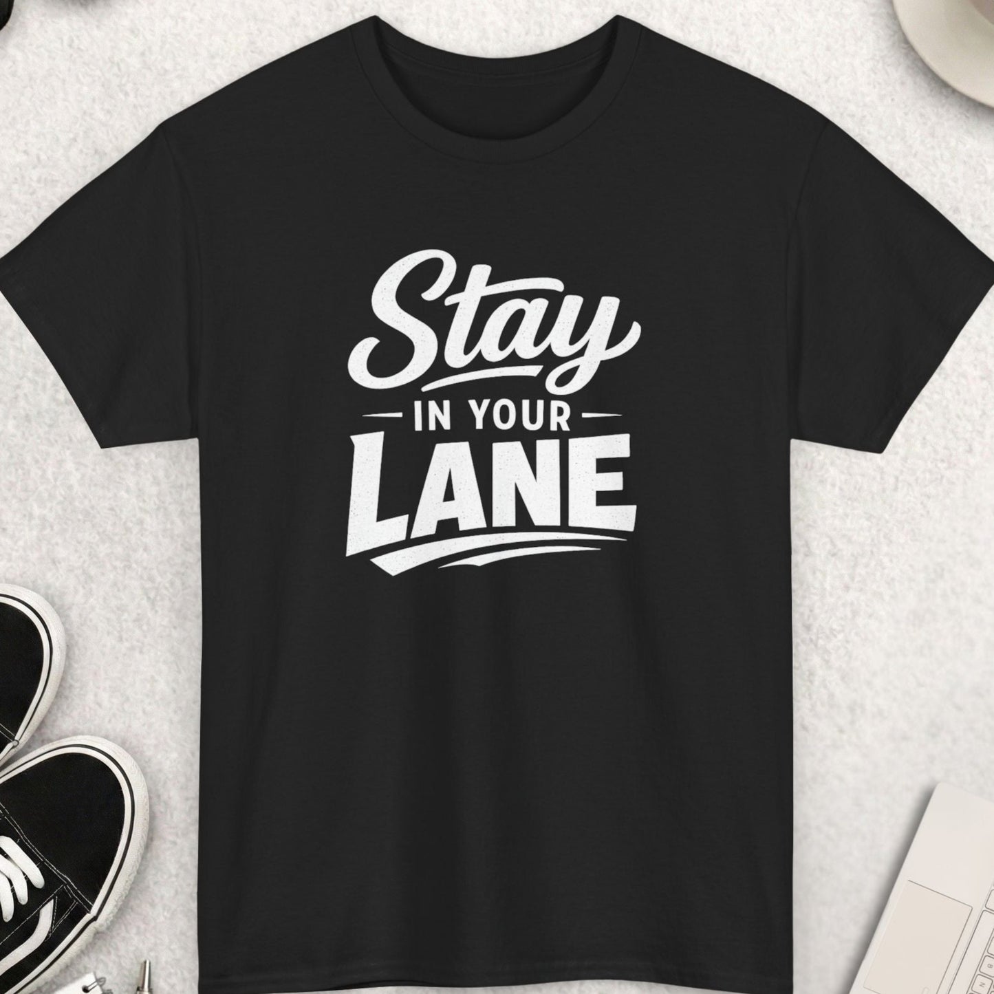 Black t-shirt with 'Stay in Your Lane' text on a light gray surface with shoes, coffee, and sunglasses.