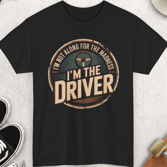 Black t-shirt with 'I'm not along for the madness, I'm the driver' design on a light gray surface with shoes, sunglasses, and a cup.