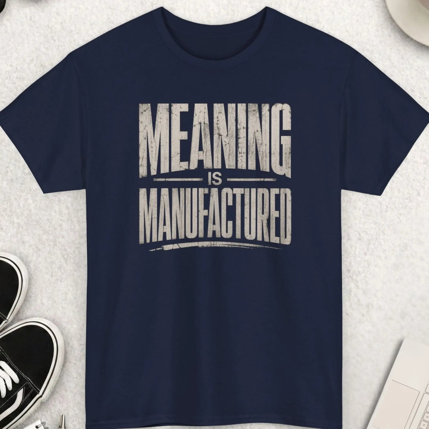 Navy blue t-shirt featuring minimalist distressed text design “Meaning Is Manufactured” in bold oversized typography
