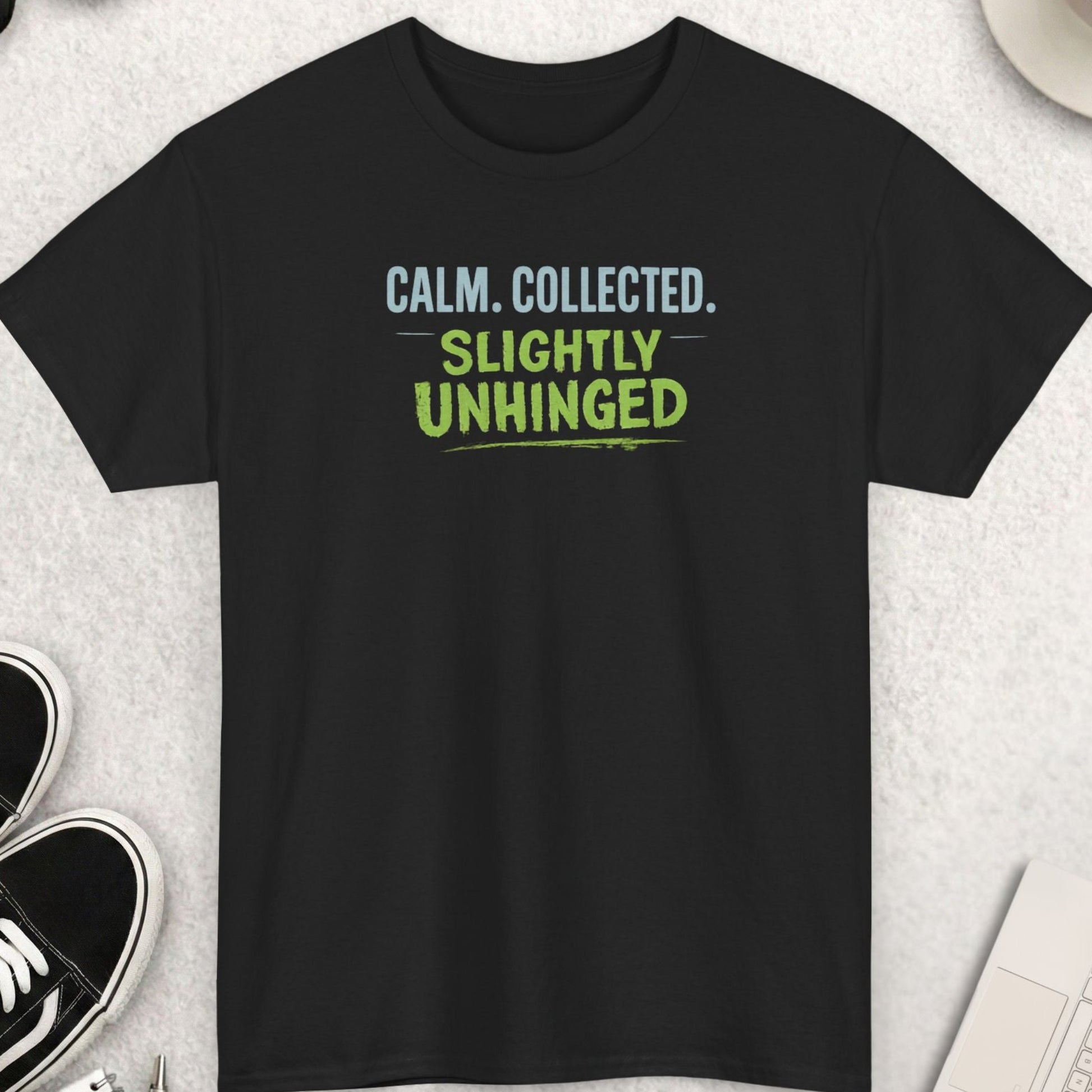 Black t-shirt with text 'CALM. COLLECTED. SLIGHTLY UNHINGED' on a light gray surface with shoes, coffee, and accessories.