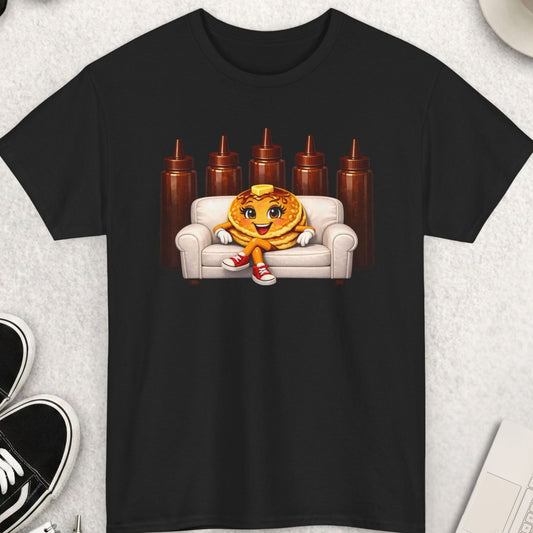 Black t-shirt with a pancake character on a couch graphic, surrounded by sunglasses, coffee, shoes, and a keyboard.