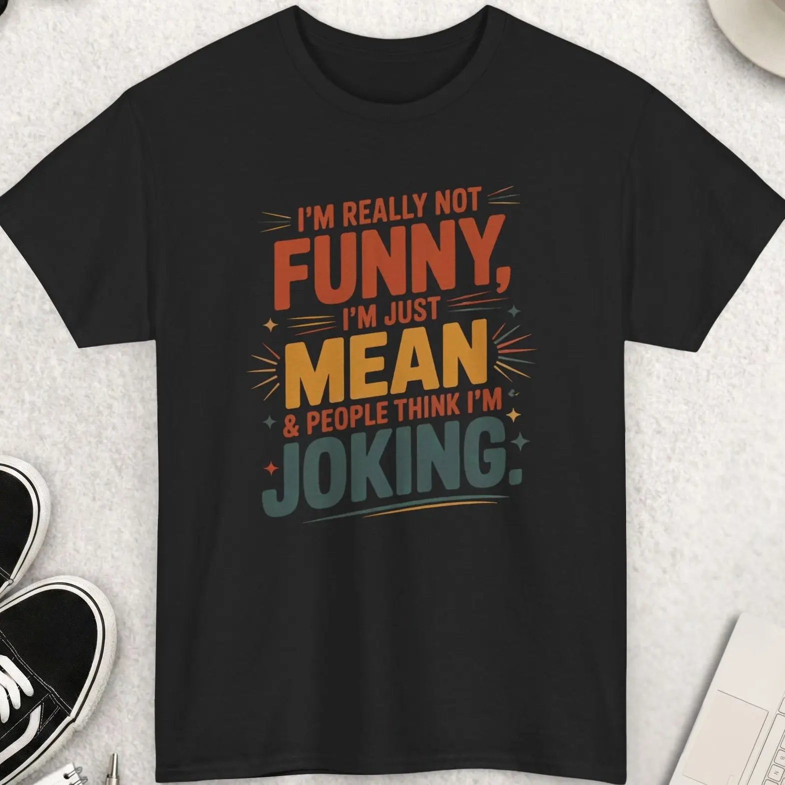 Black t-shirt with humorous text on a light gray surface with shoes, coffee, and sunglasses.