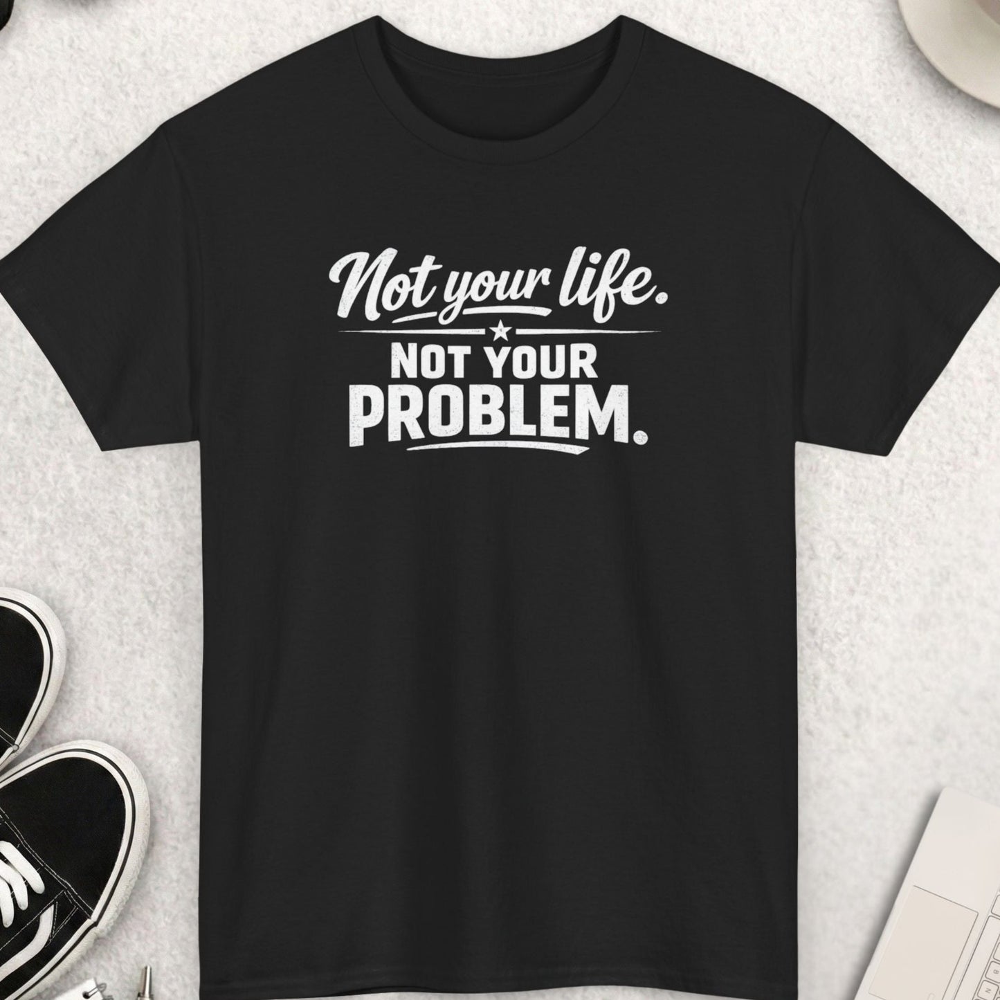 Black t-shirt with 'Not your life. Not your problem.' text on a light gray surface with shoes, coffee, and accessories.